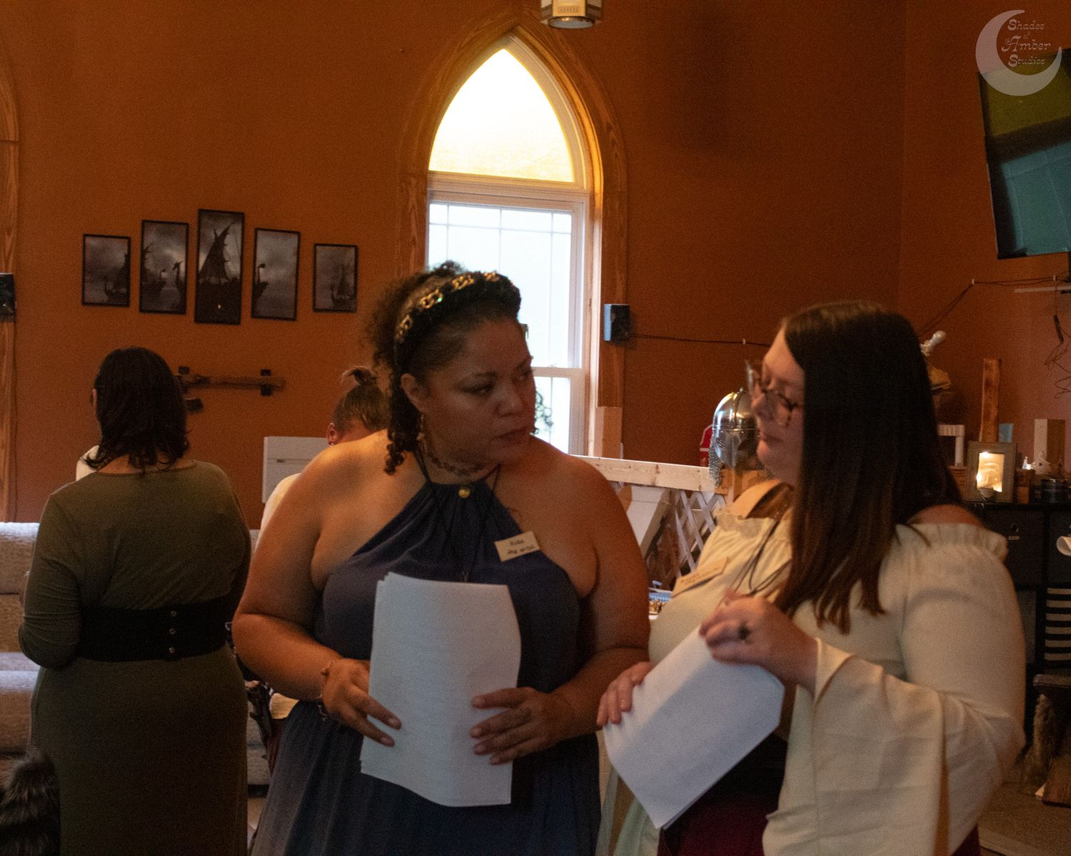 Two people in formal attire converse while reviewing documents inside a room with arched windows and orange walls.