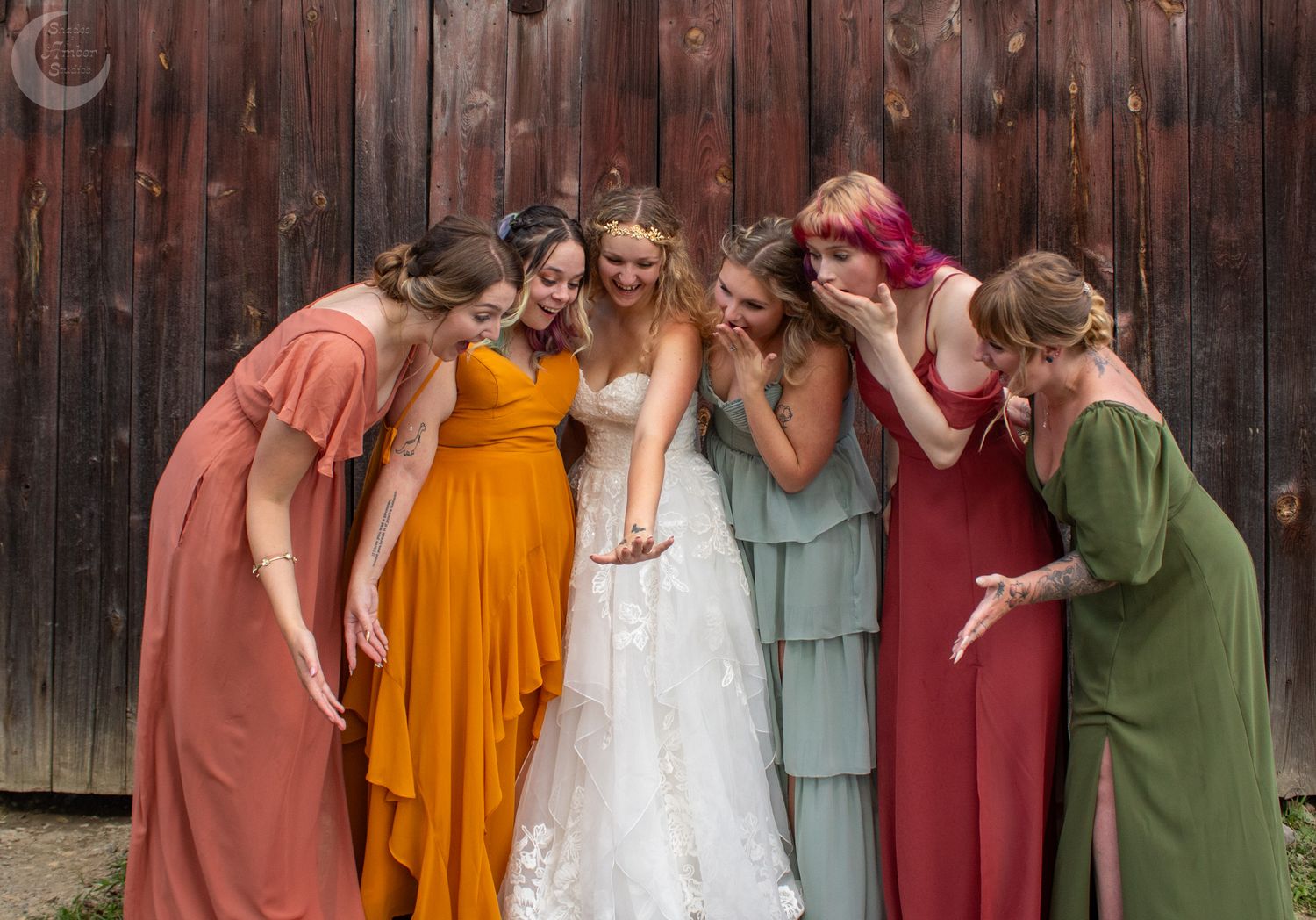 A bride and her bridesmaids in colorful autumn dresses share a lighthearted moment against a rustic wooden wall.