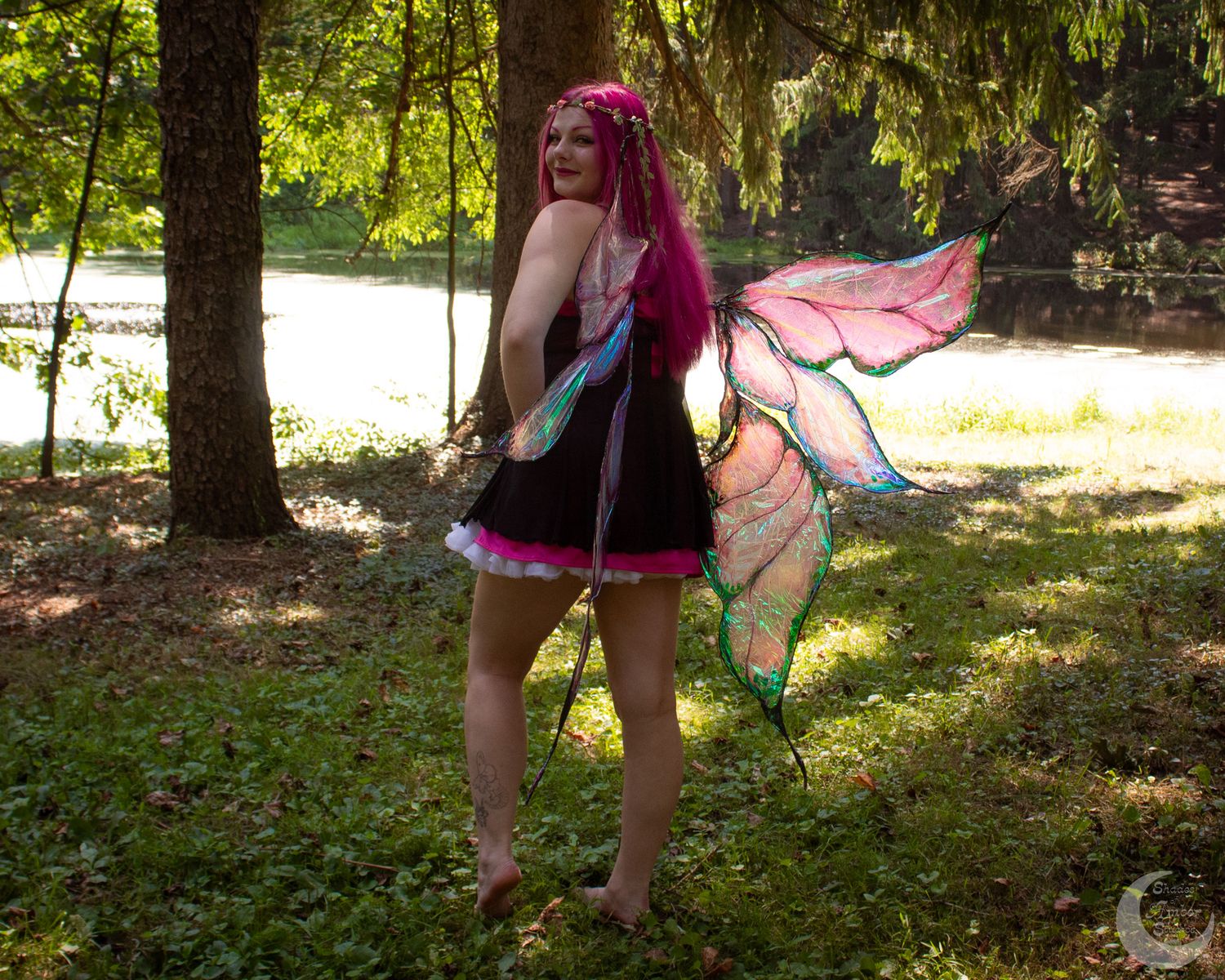 A person in a black costume and pink fairy wings performs an aerial dance routine in a lush green forest setting.