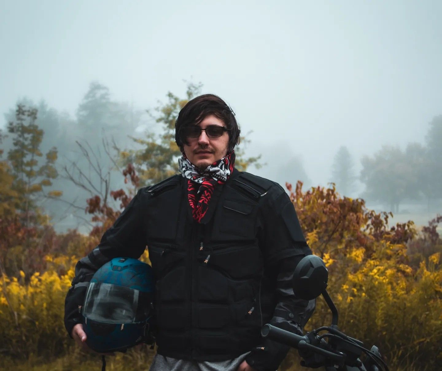 Motorcyclist in black jacket and bandana standing with bike against foggy fall landscape.