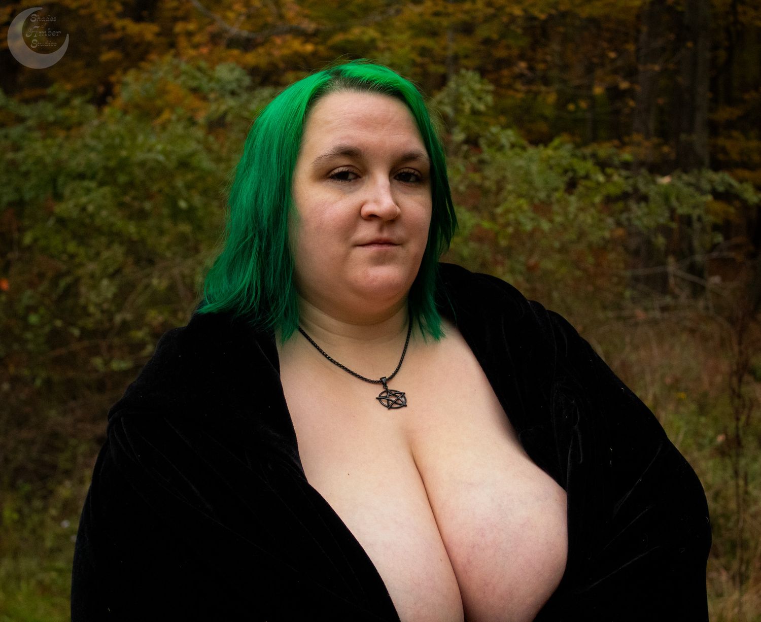 Plus size model with vibrant green hair poses in black cape against fall foliage.