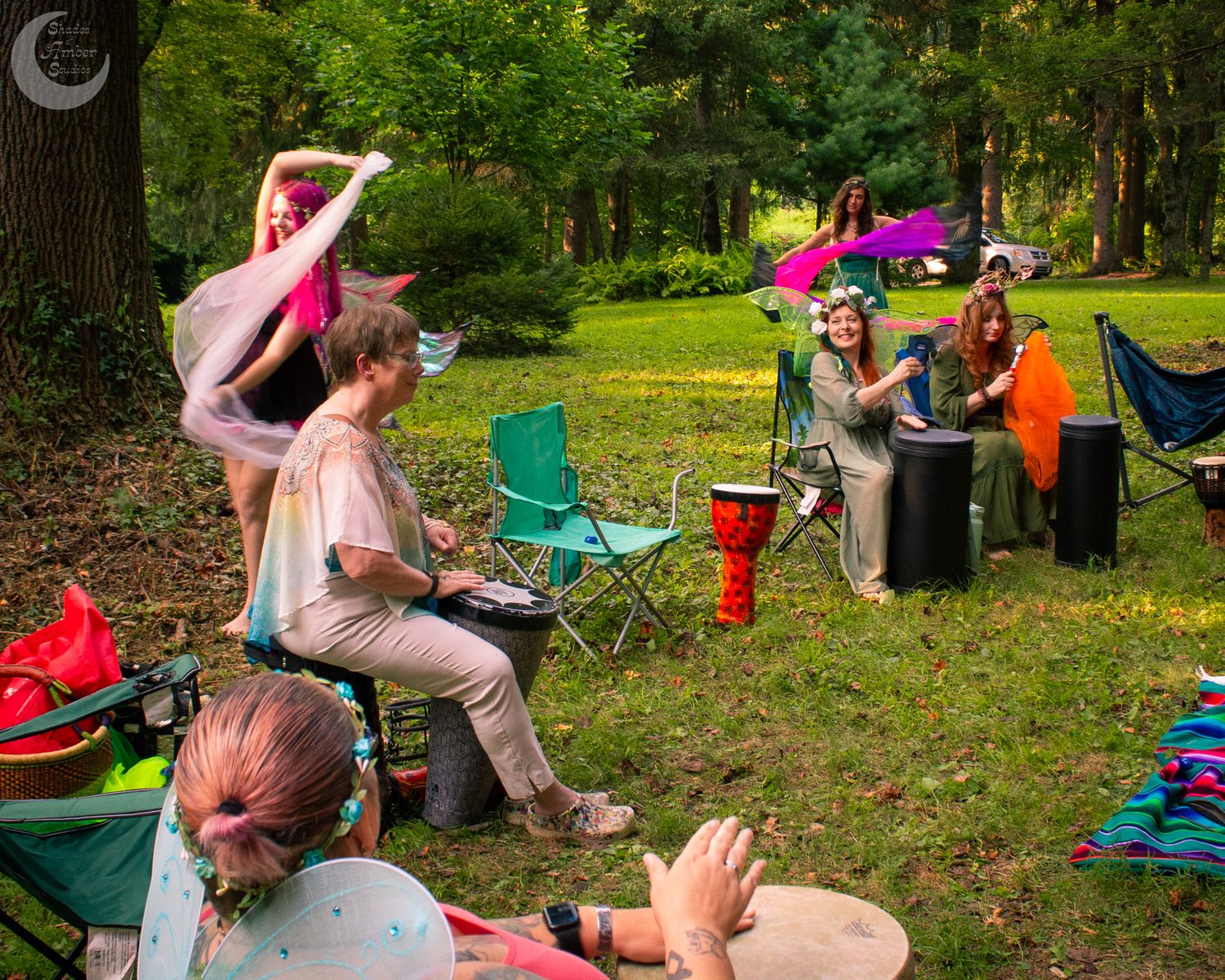 Colorful outdoor gathering in a wooded area with camping chairs and musical instruments.