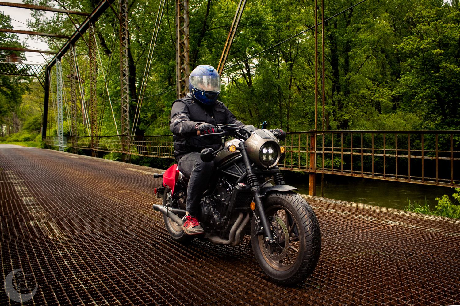 A motorcycle rider crosses a rustic metal bridge on a black custom motorcycle in a lush forest setting.