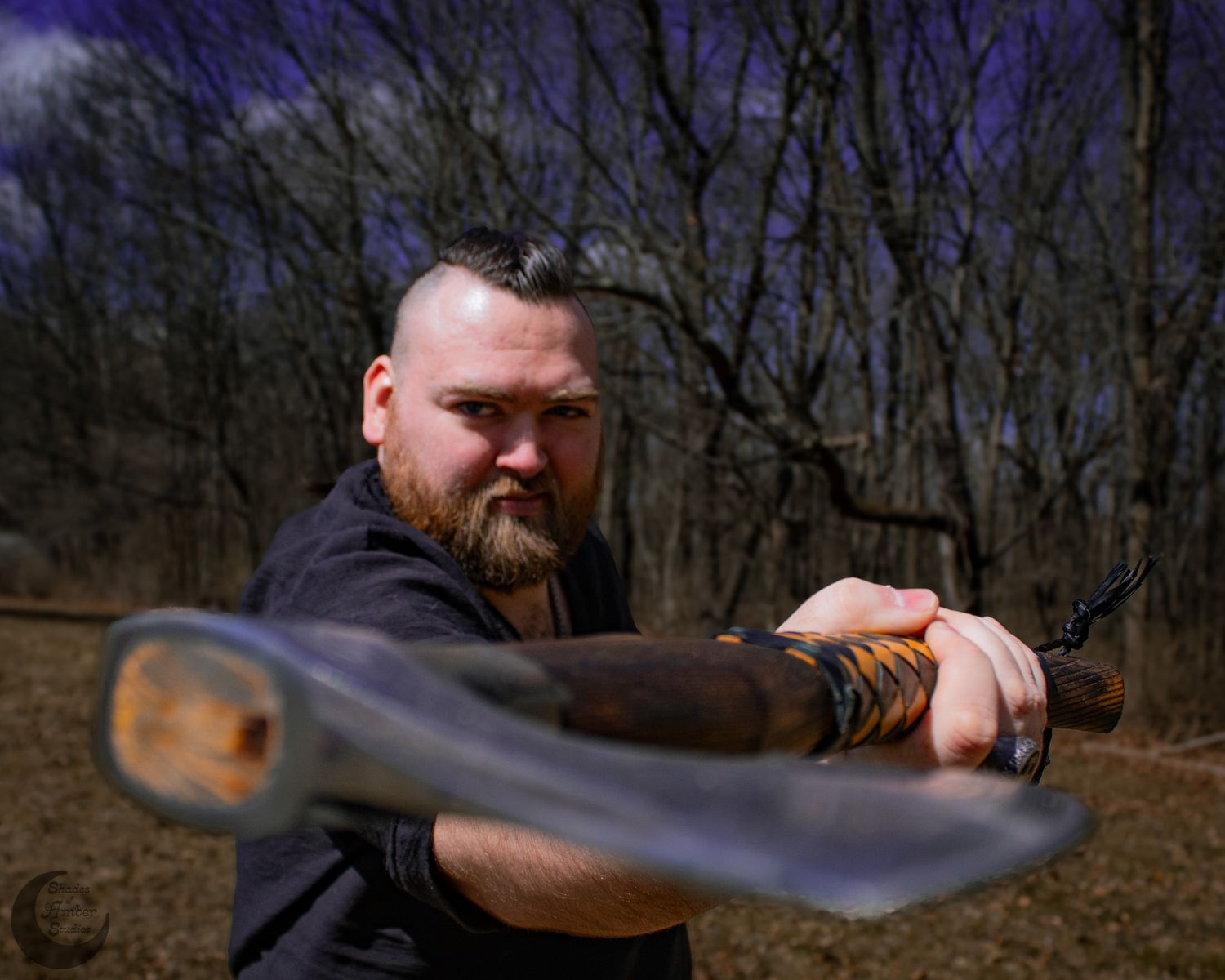 A bearded man with a mohawk wields a large sword outdoors in a wooded area, posing in an aggressive stance.