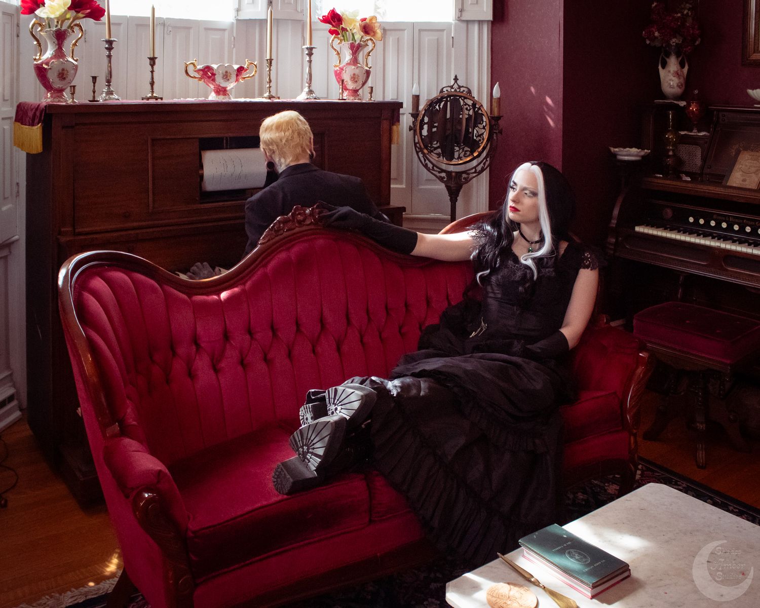 Two women in gothic black dresses pose in a Victorian parlor with red velvet sofa and antique piano.