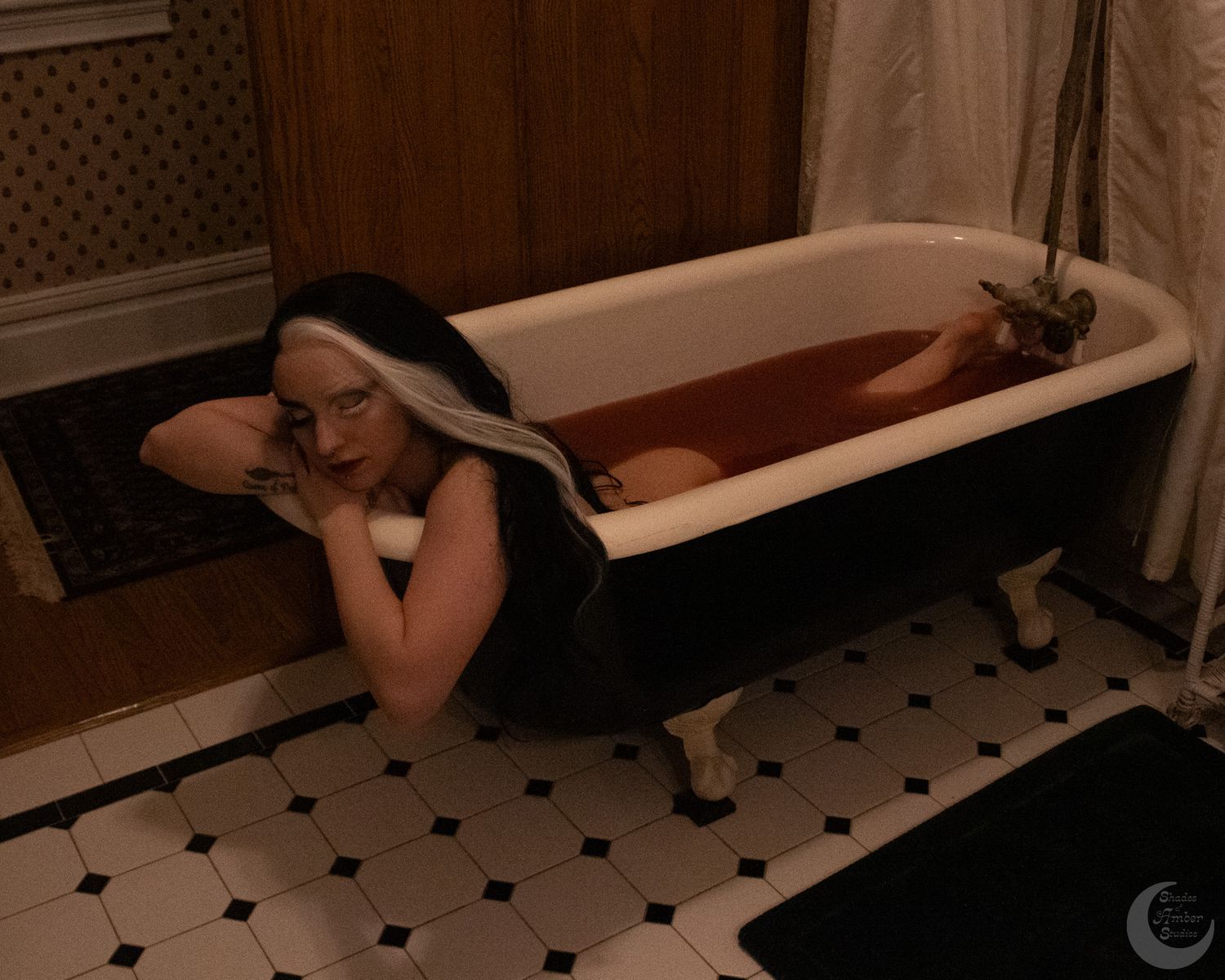 A woman with black and white hair relaxes in a vintage clawfoot bathtub filled with red water in a tiled bathroom.