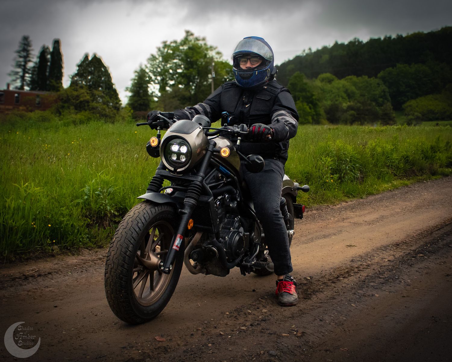 A motorcycle rider in protective gear sits on a black custom bike on a dirt road with green fields in the background.