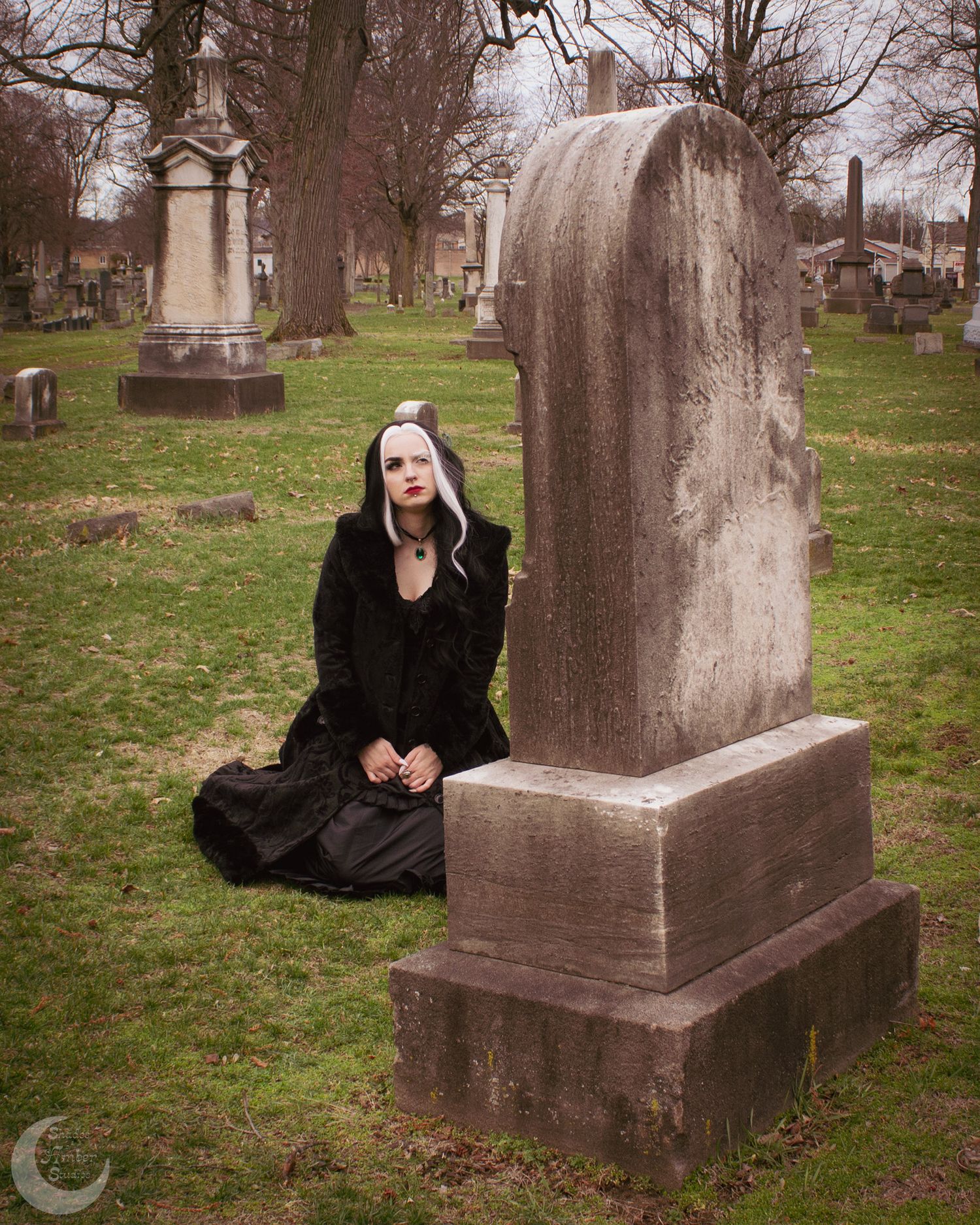 Gothic woman in black dress kneeling beside a large gravestone in a cemetery.