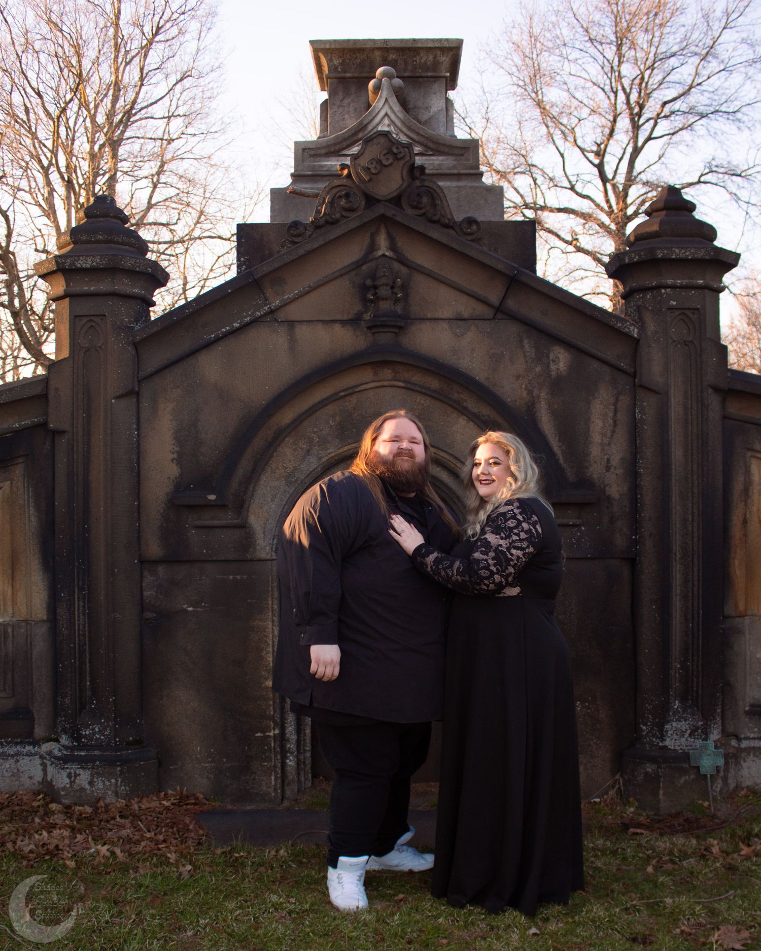 Couple in black gothic outfits posing in front of ornate stone archway gate in outdoor cemetery setting.