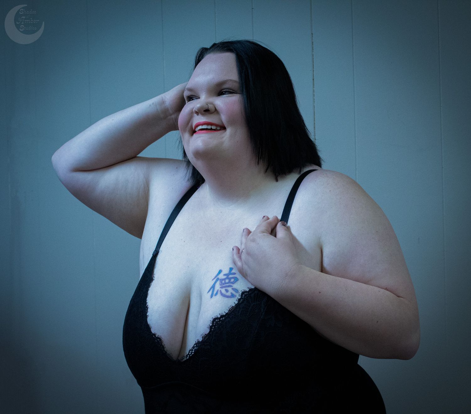 A joyful person in a black tank top poses against a blue-tinted wall with one hand behind their head.