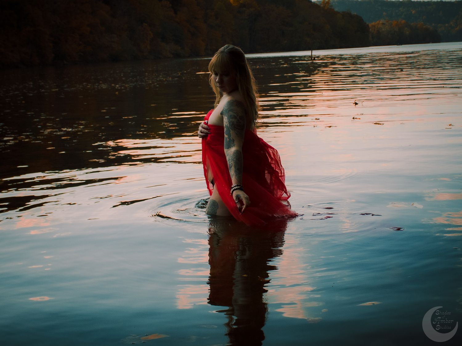 Figure in red flowing dress standing in calm water during sunset with reflective surface.
