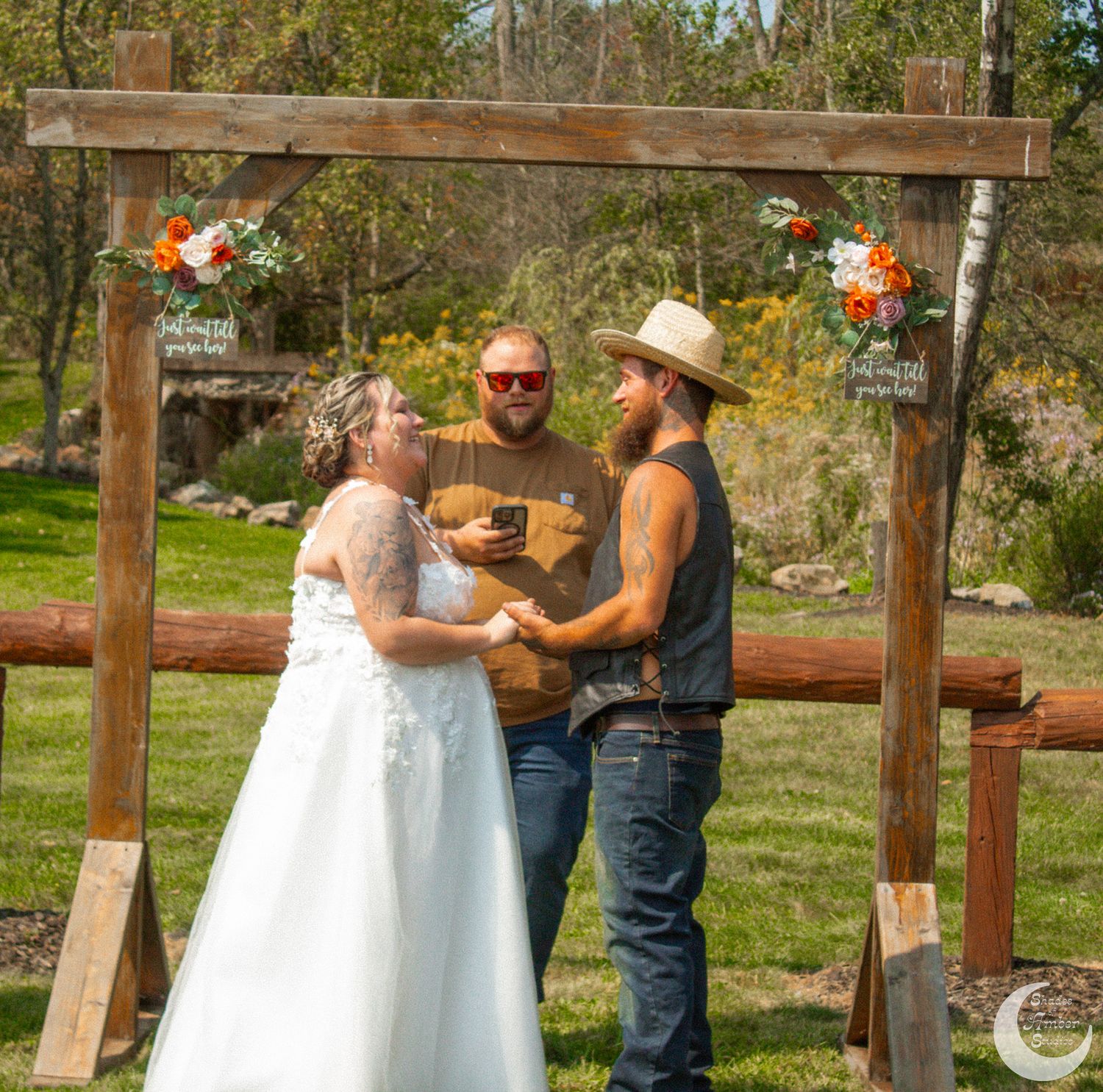 Wedding ceremony taking place under wooden arch decorated with flowers in outdoor rustic setting.