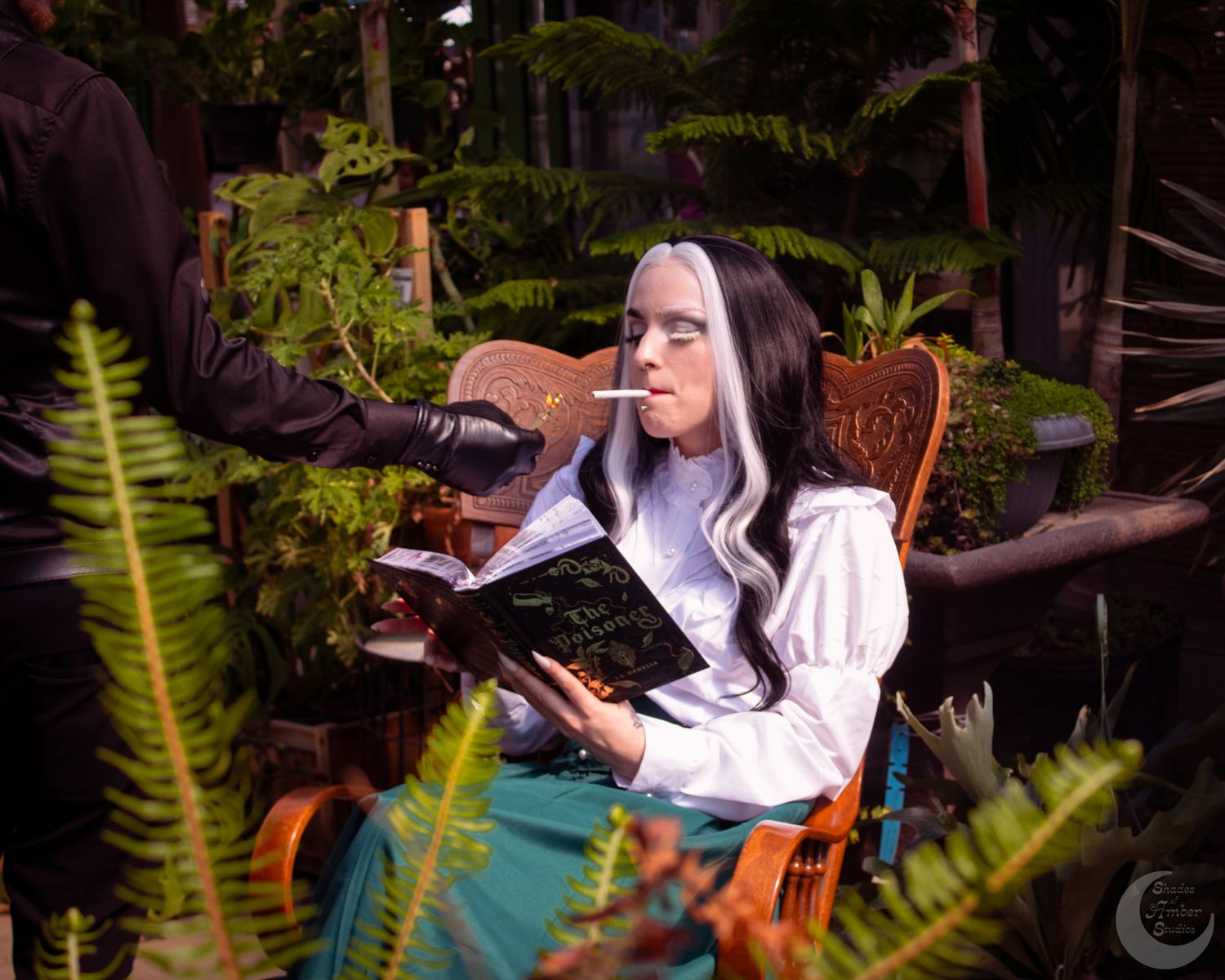 Woman cosplaying with white hair, reading a black book while seated among lush green plants in a garden setting.