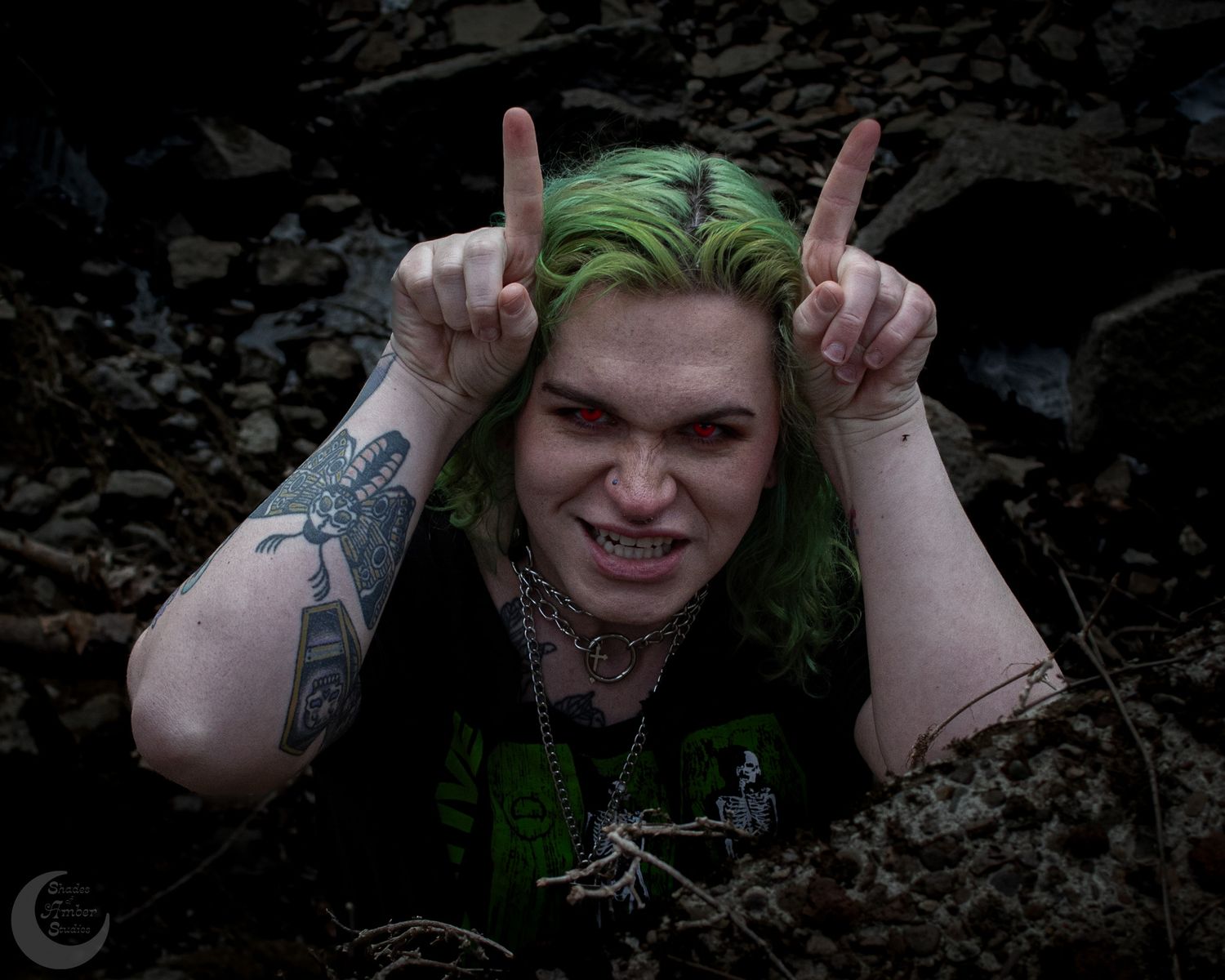 Alternative model posing with green hair making horns with fingers while emerging from dark background.