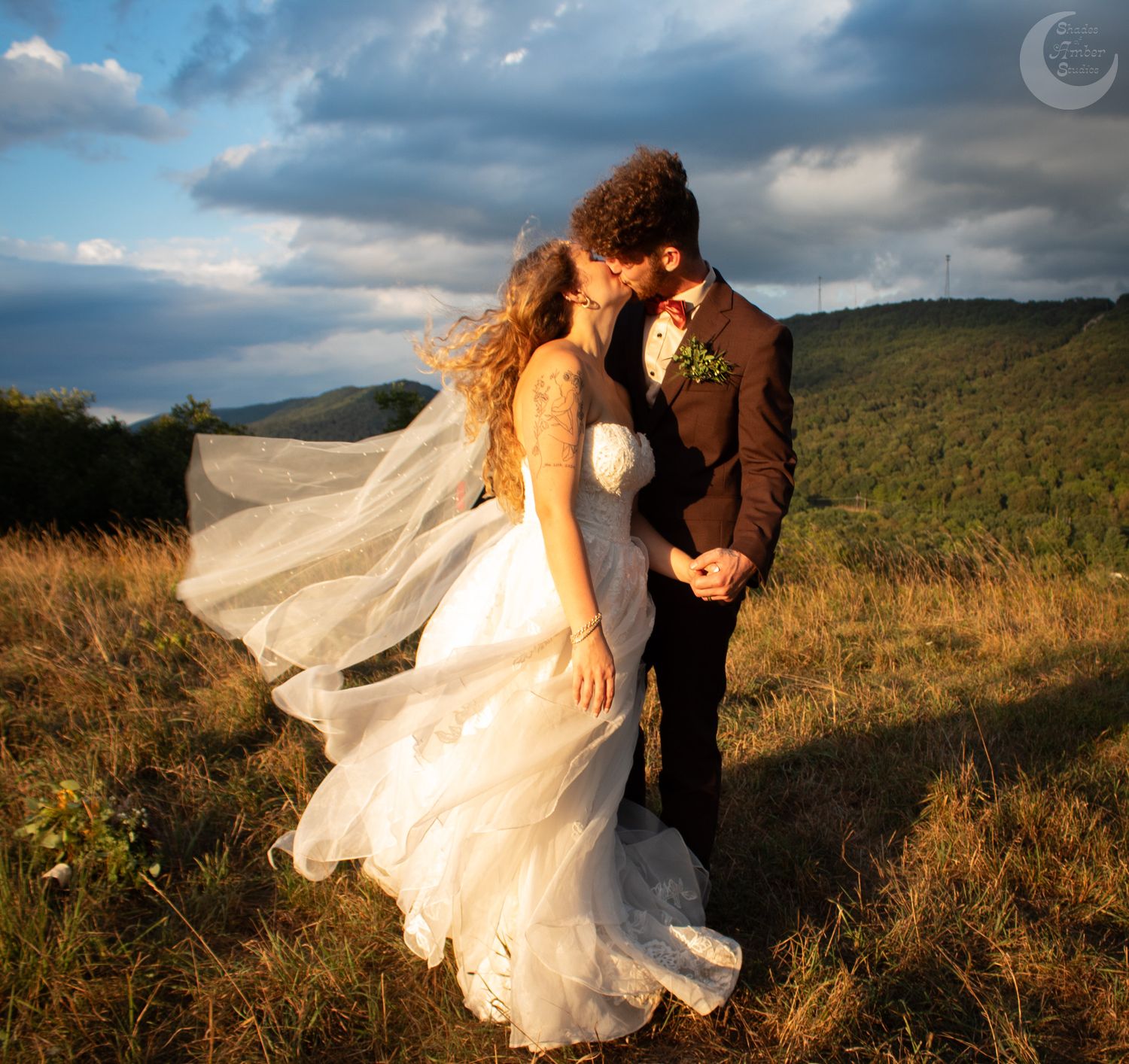 A couple in wedding attire dances in a scenic mountain field at sunset as the bride's dress and veil flow in the wind.