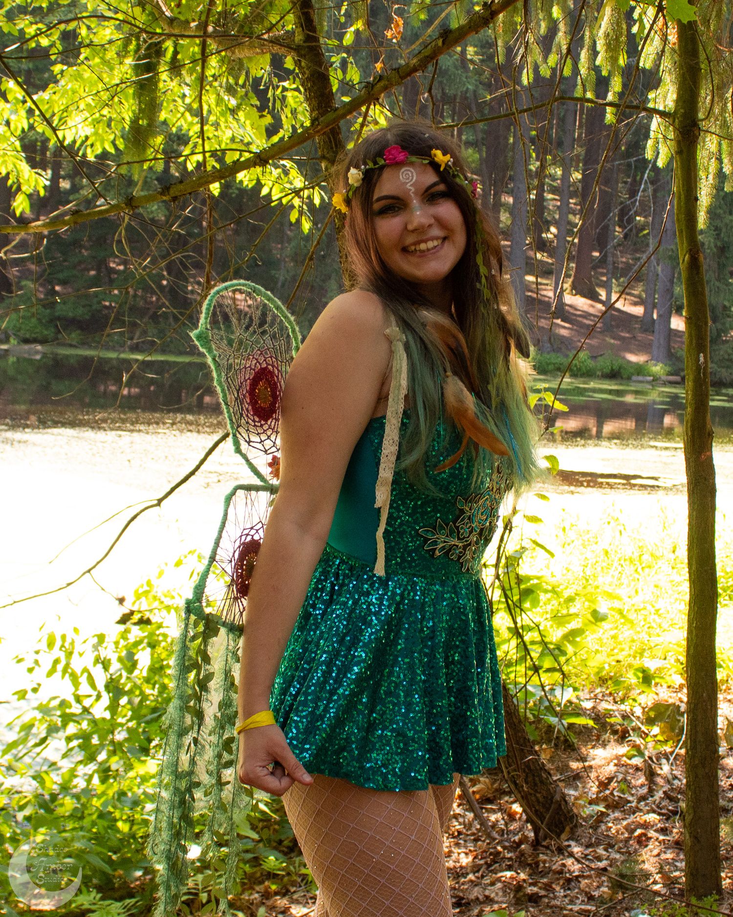 A person poses in a shimmering green sequin dress among trees in a sunlit outdoor setting.