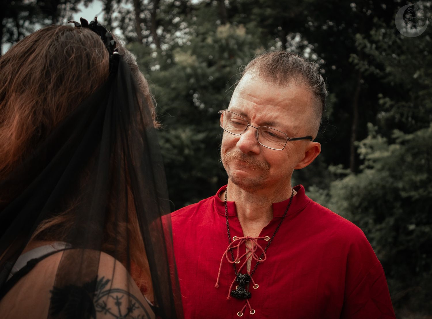 A person wearing a red shirt and glasses speaks closely with someone in dark hair against a forested background.