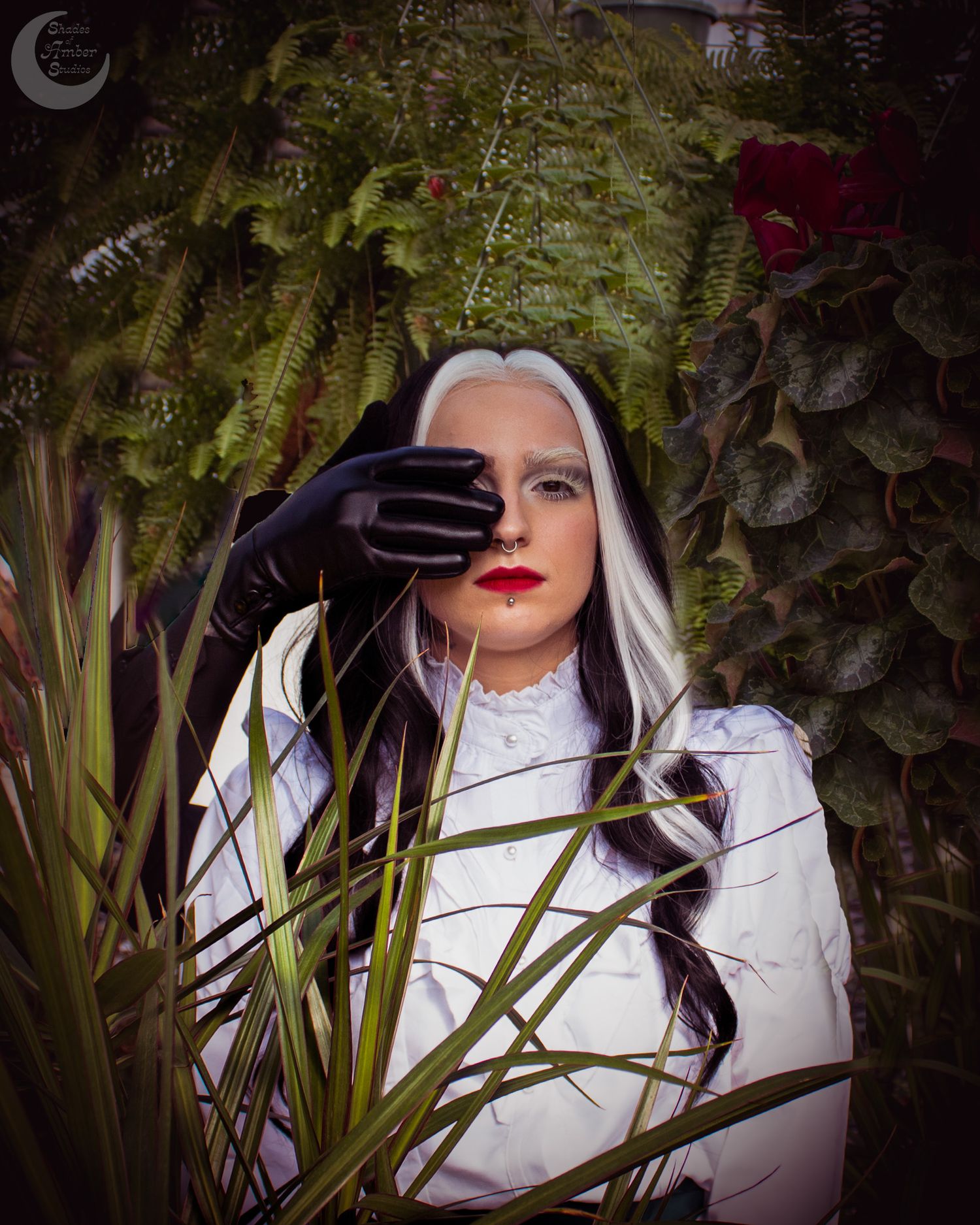 Woman in white dress and black gloves with gothic makeup poses among lush green ferns and red roses.