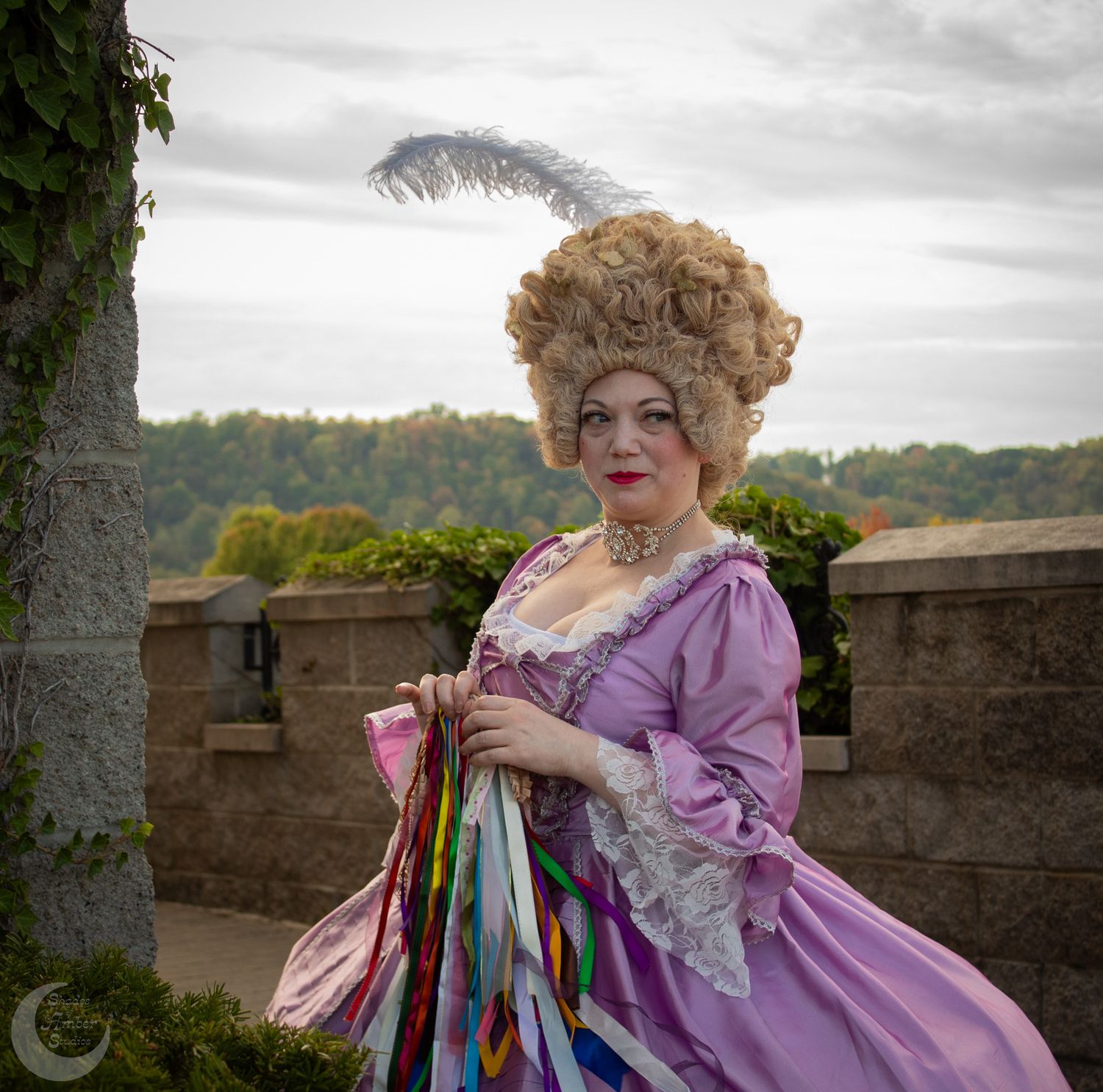 A period costume featuring a lavender dress and elaborate wig with feather against a stone wall.