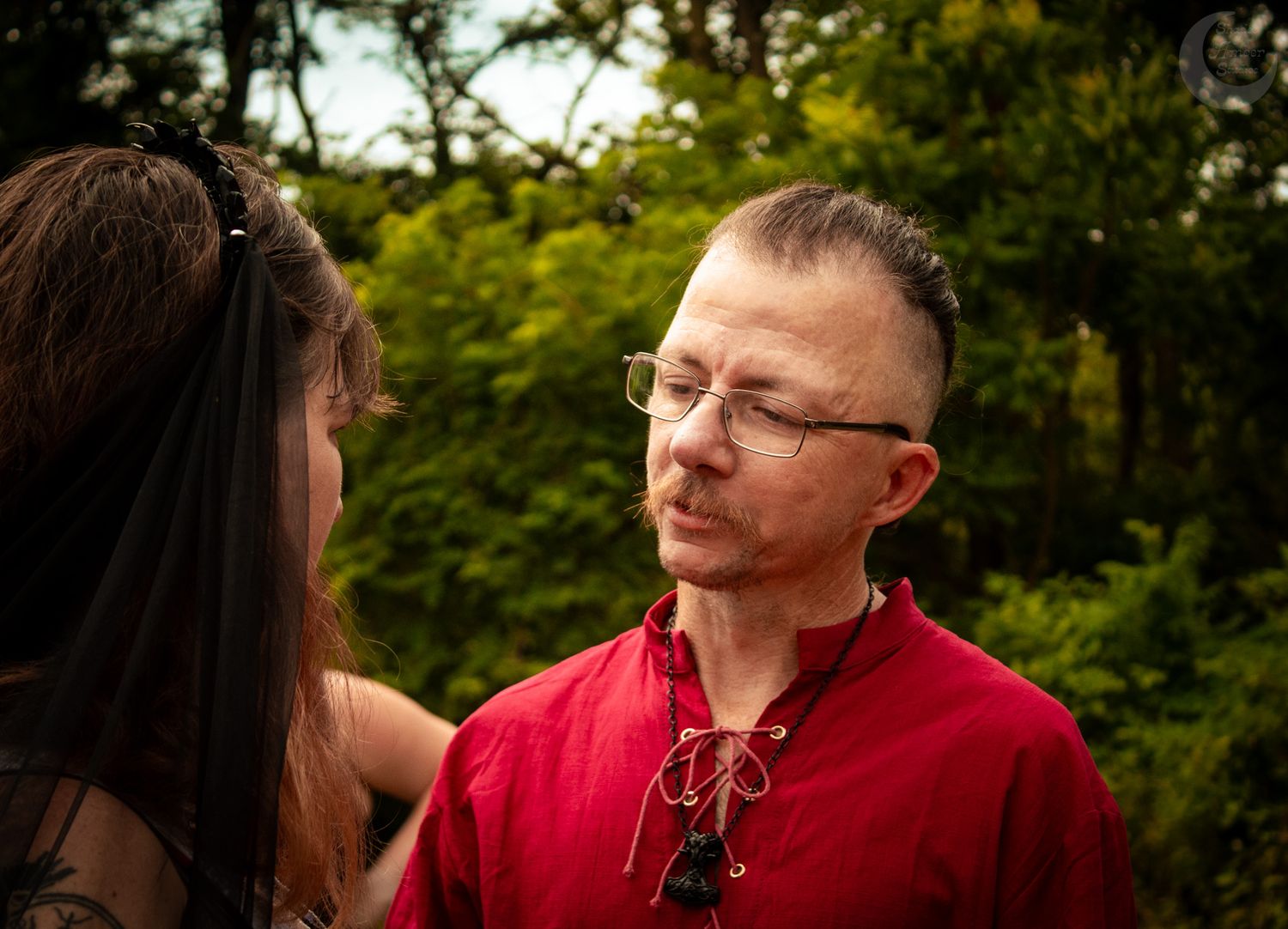 Two people in conversation outdoors against a green leafy background, one wearing a bright red shirt.