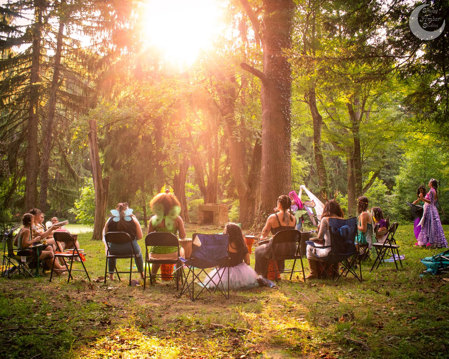 People gather in camping chairs among tall trees at sunset in a peaceful outdoor setting, creating a warm golden atmosphere.