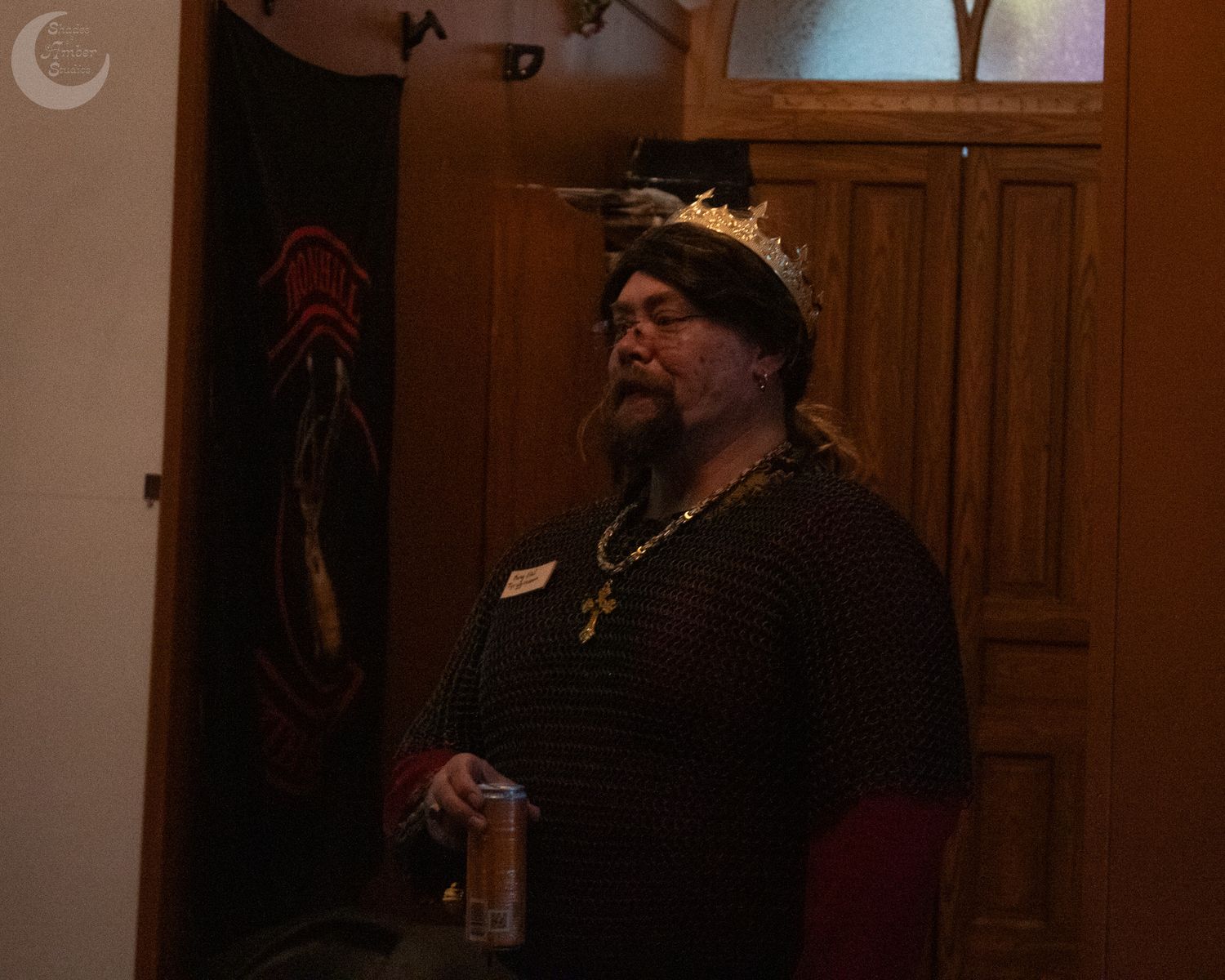 A figure wearing medieval attire including a crown and dark clothing stands in dim lighting indoors.