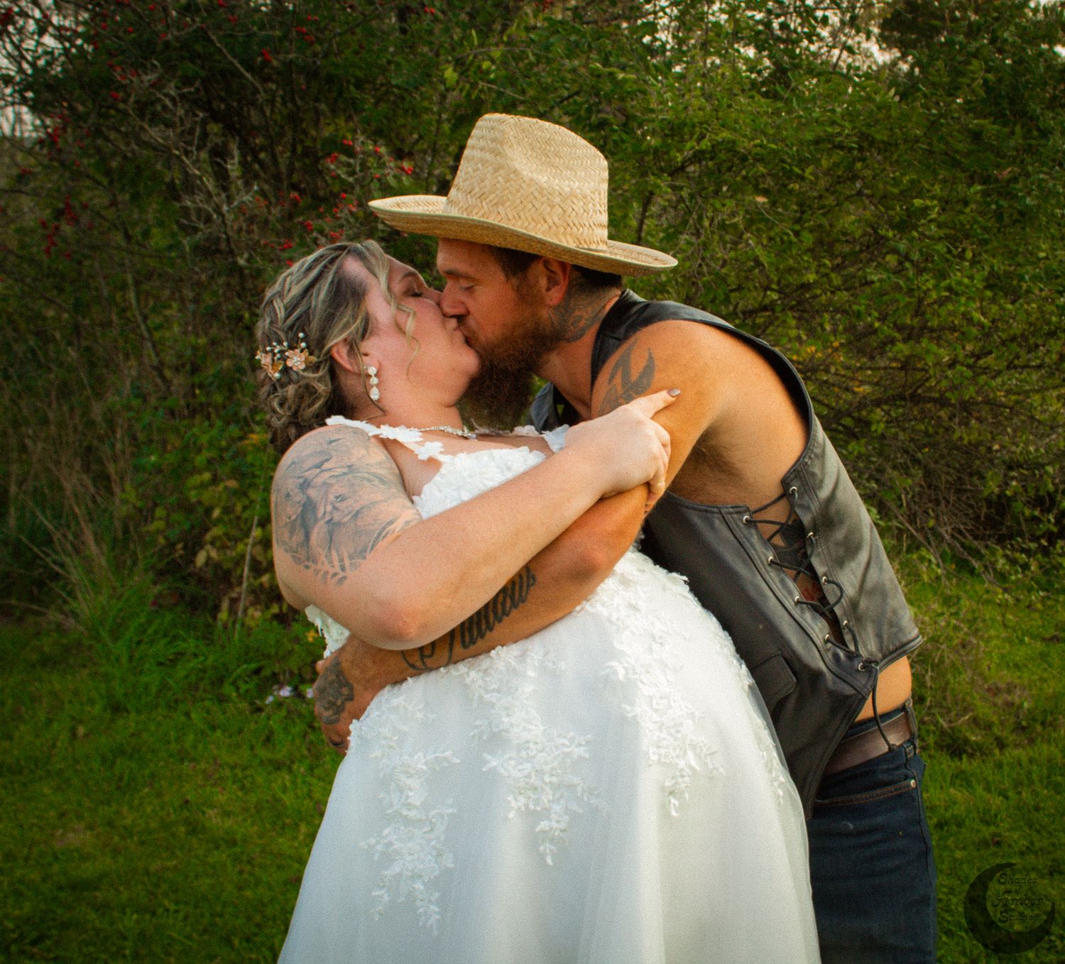 A romantic wedding photo outdoors showing a tender kiss between a couple in rustic attire with greenery background.