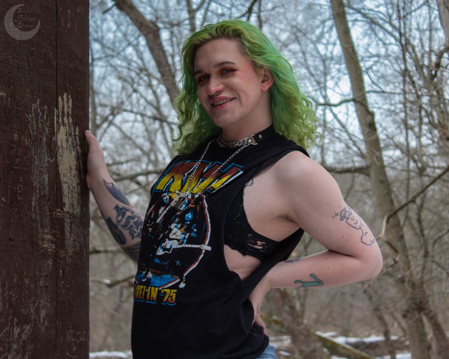 Figure in sleeveless band shirt with bright green hair poses against tree in winter forest setting.