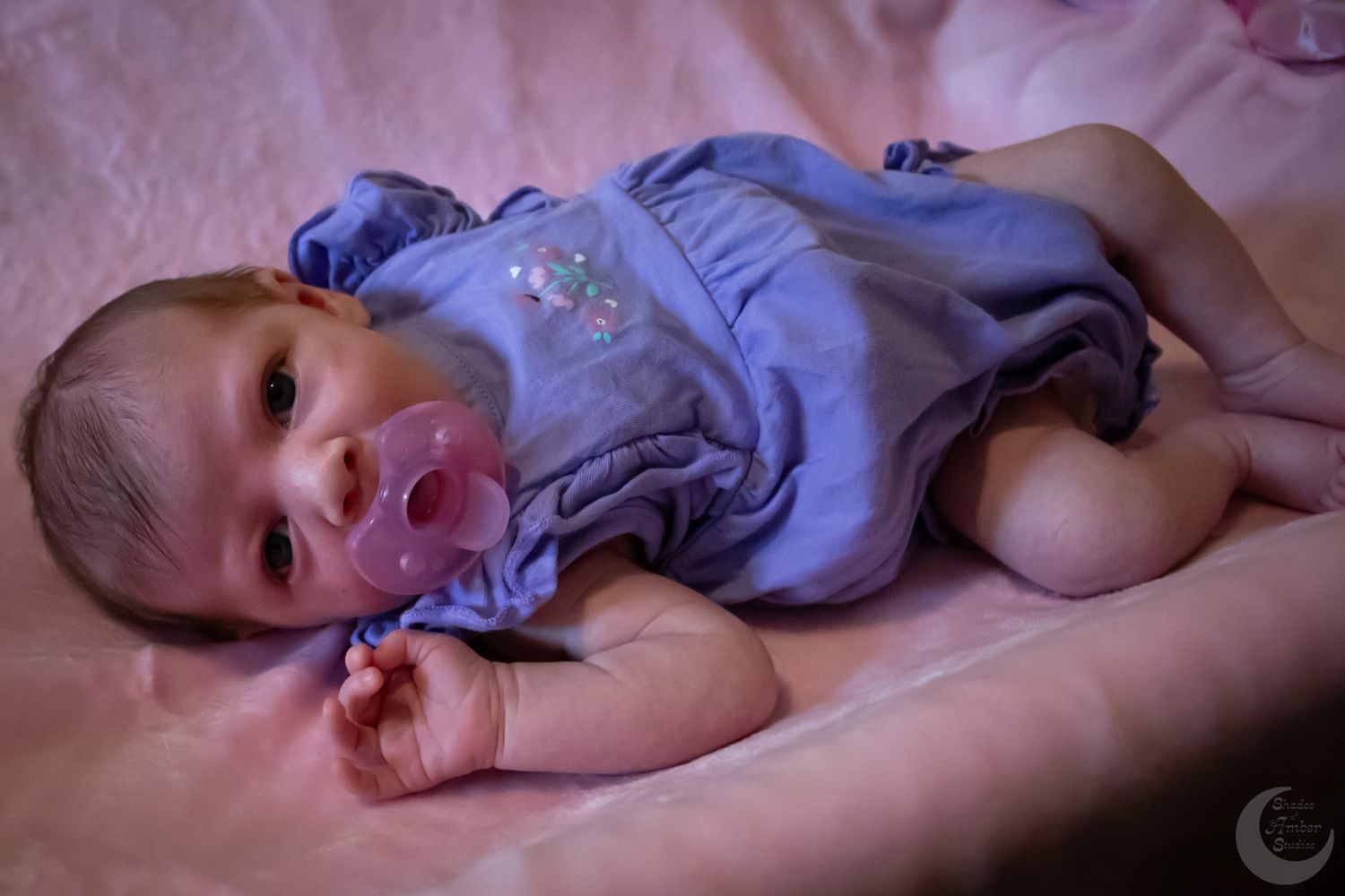 A baby wearing a purple onesie lies on pink bedding with a sweet expression.