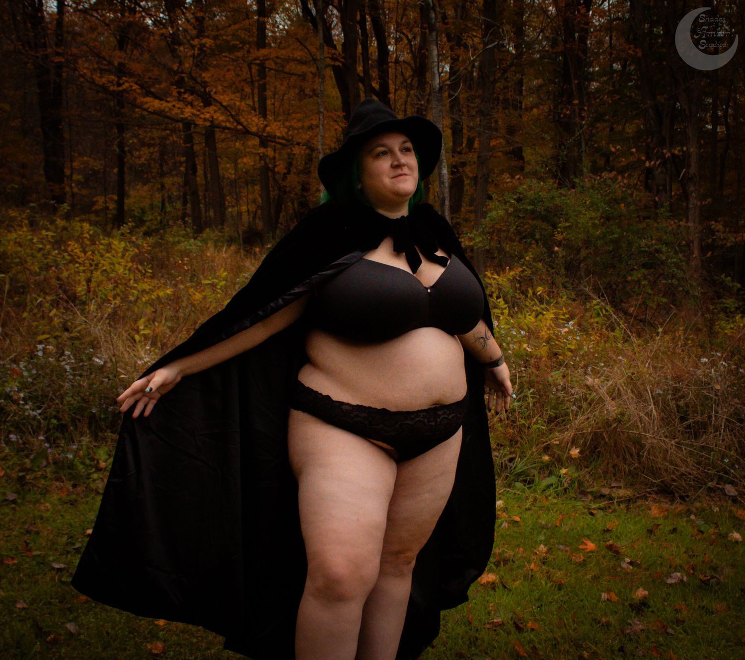Person wearing black cape and lingerie poses confidently in autumnal woods.