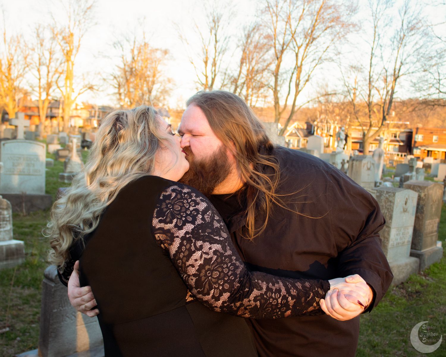 Couple sharing a romantic kiss at golden hour in a historic cemetery with gravestones behind.