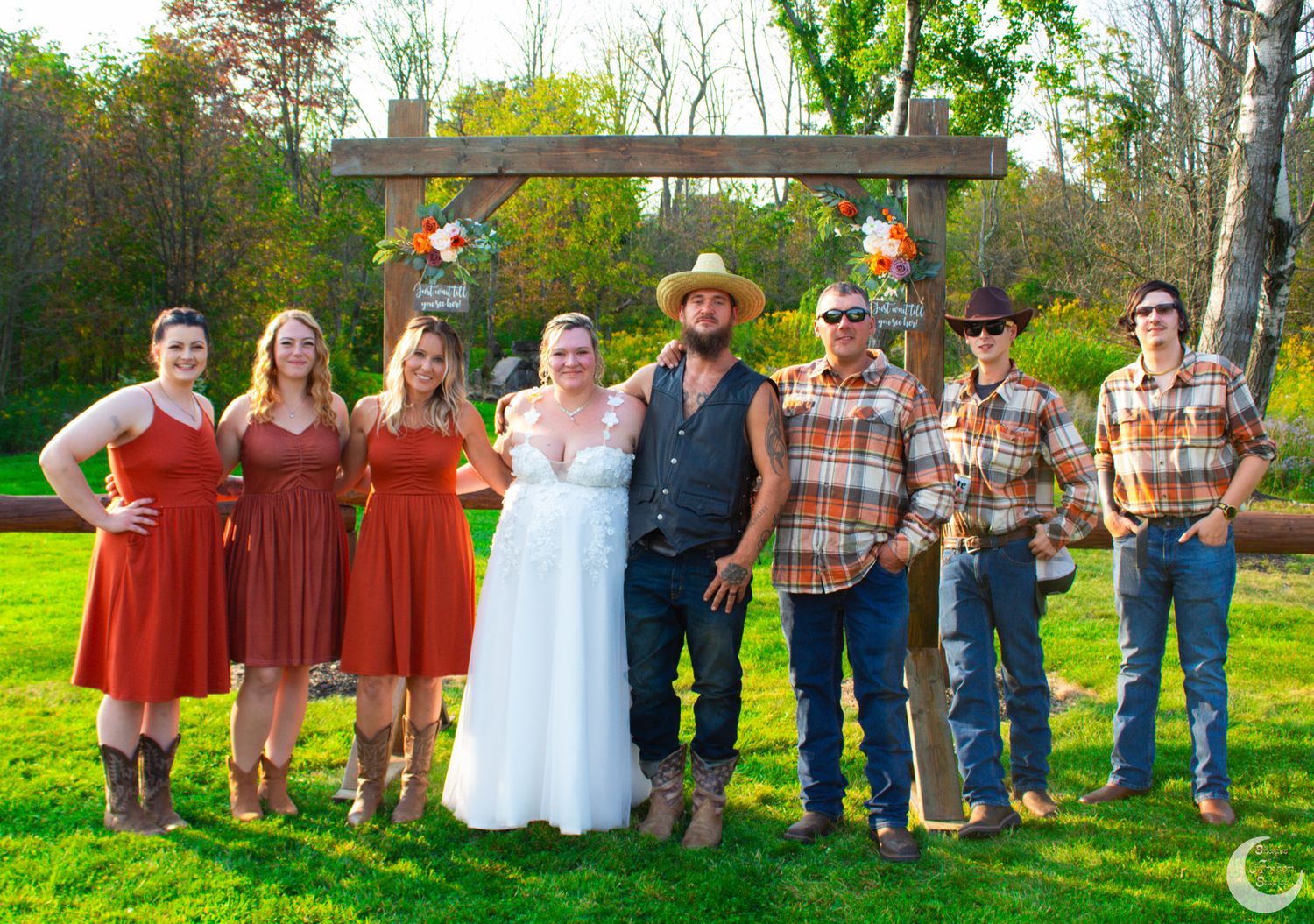 Wedding party in burnt orange dresses and plaid shirts posed under wooden arch at outdoor ceremony.