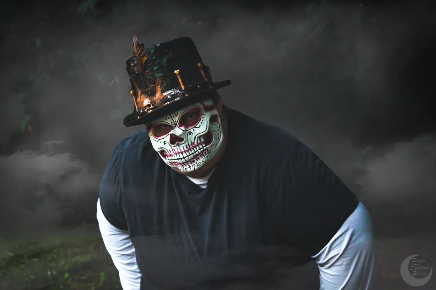 Spooky figure in black wearing decorated skull mask and hat against dark cloudy background.