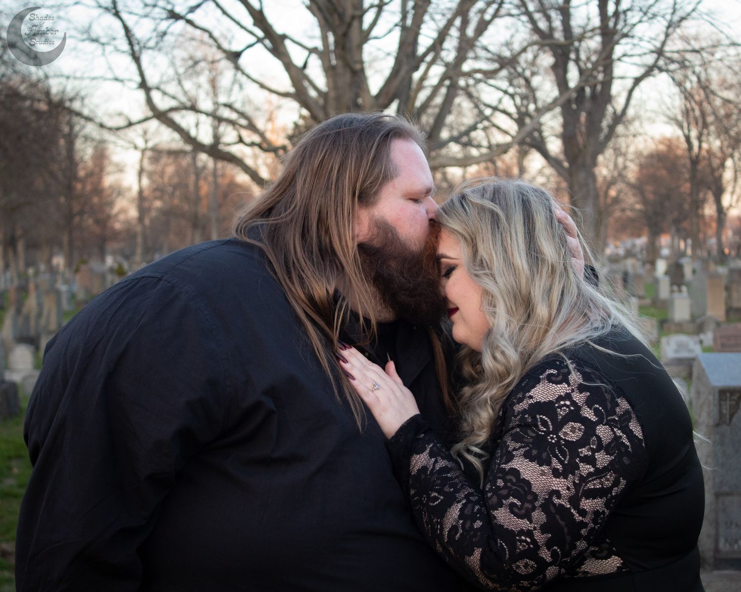 Couple sharing a romantic kiss in a cemetery at dusk, both dressed in black lace and formal attire.