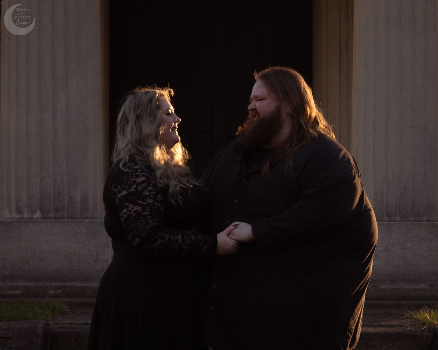 A couple dressed in black holding hands and smiling at each other in front of a grand building entrance.