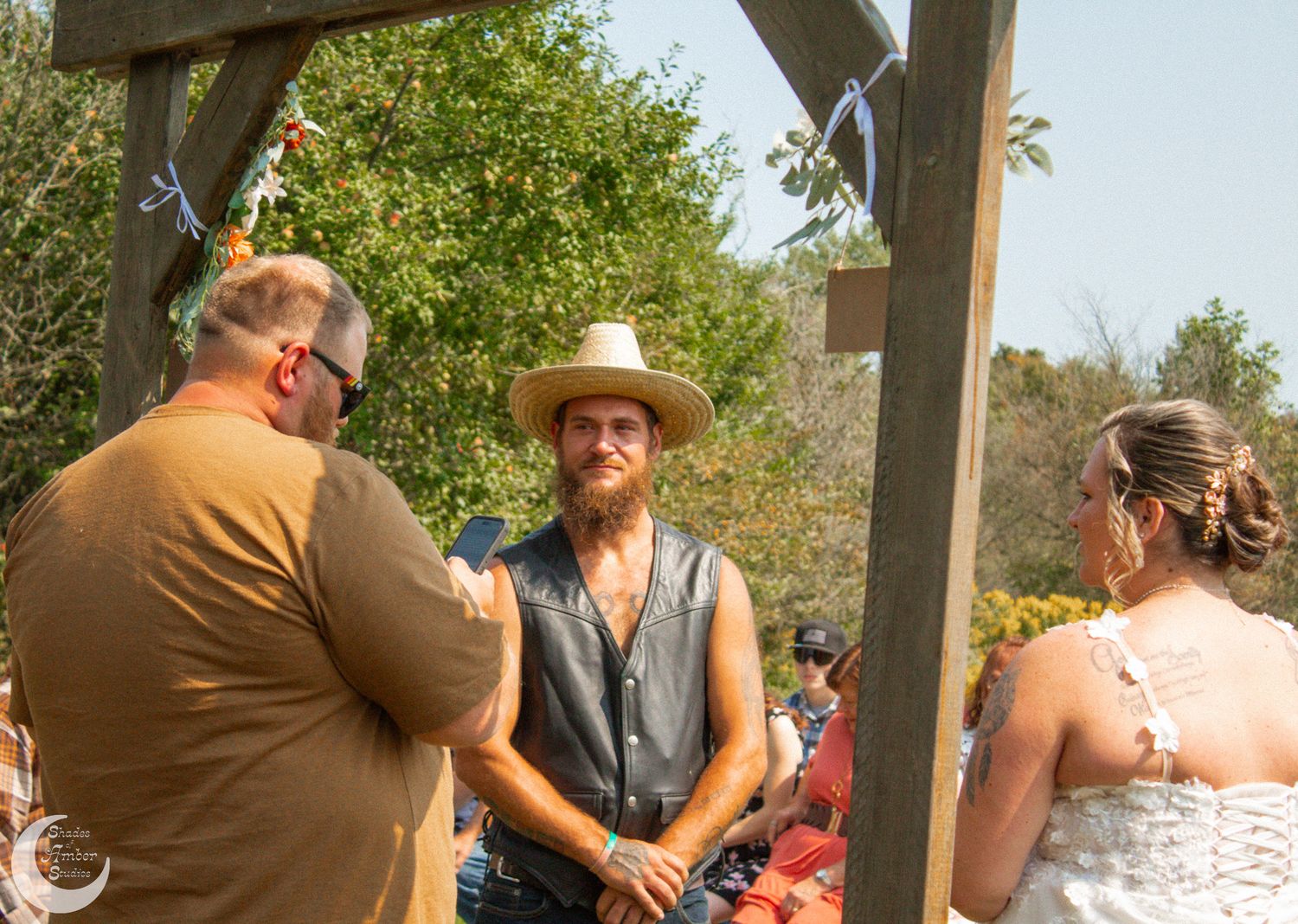Outdoor wedding ceremony under wooden arbor with guests and scenic countryside views.