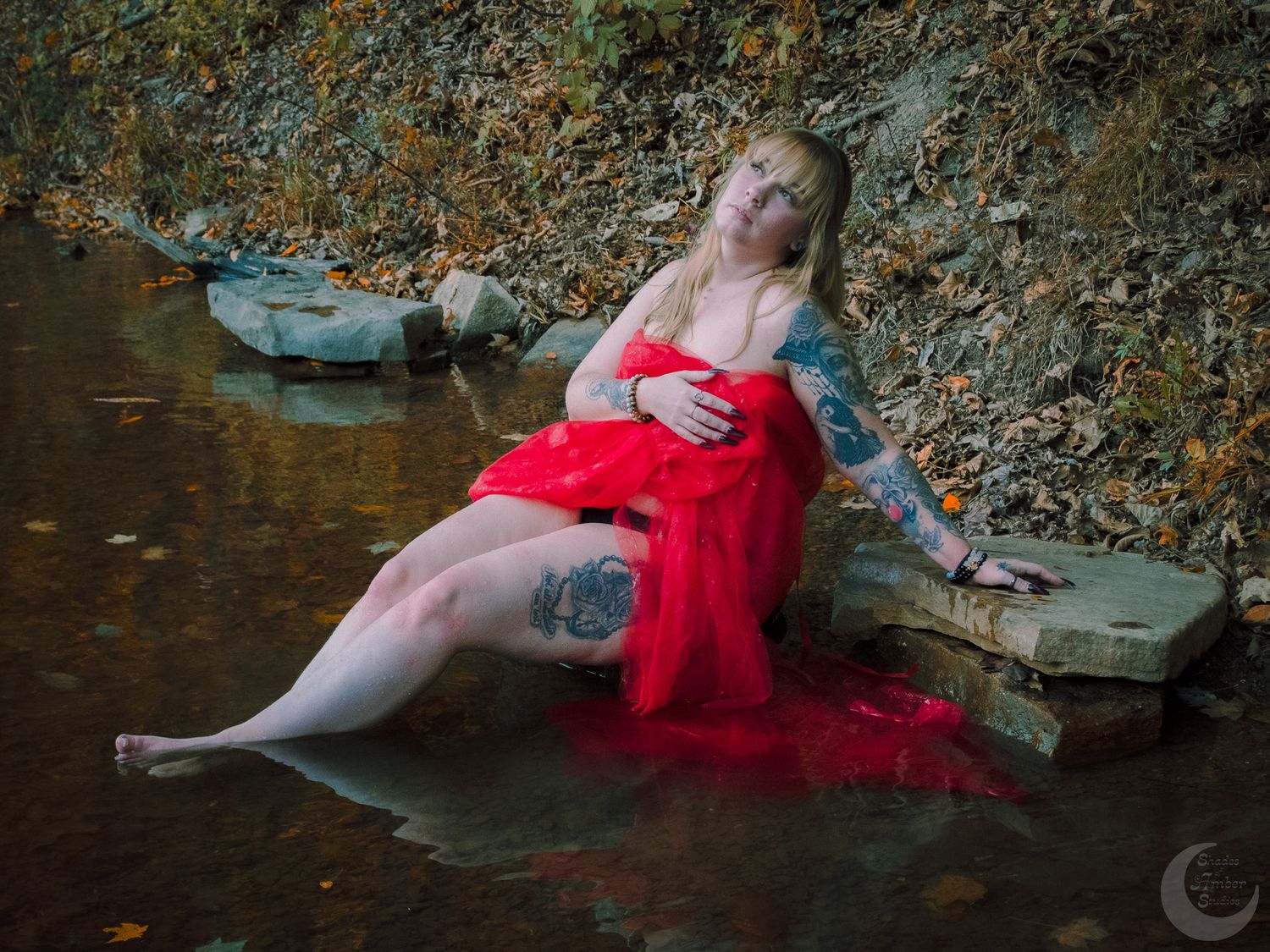 Dreamy portrait of person in flowing red fabric reclining by water's edge in autumn setting.