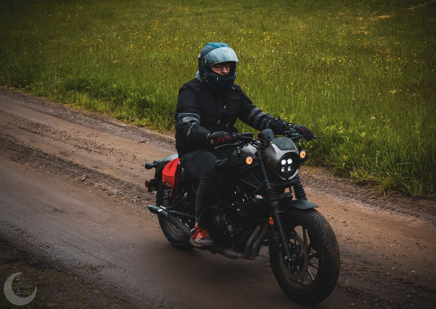 A motorcyclist in black gear rides a dark sport bike on a dirt road with green grass in the background.