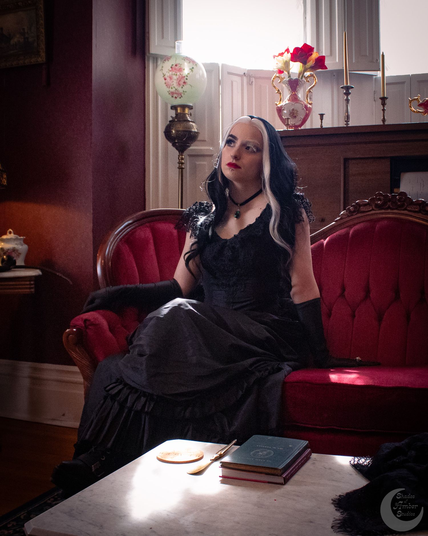 Woman in gothic black dress with white-streaked hair poses on red velvet sofa in Victorian-style room.