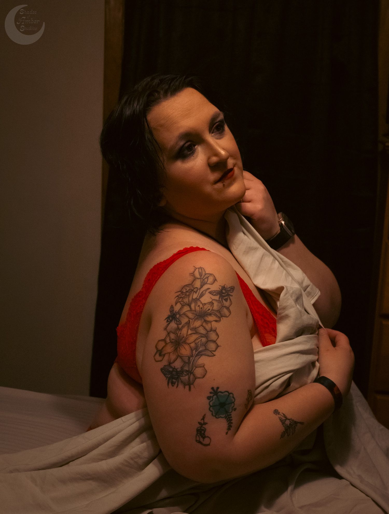 Woman with floral arm tattoos poses thoughtfully in red and white outfit in dimly lit room.