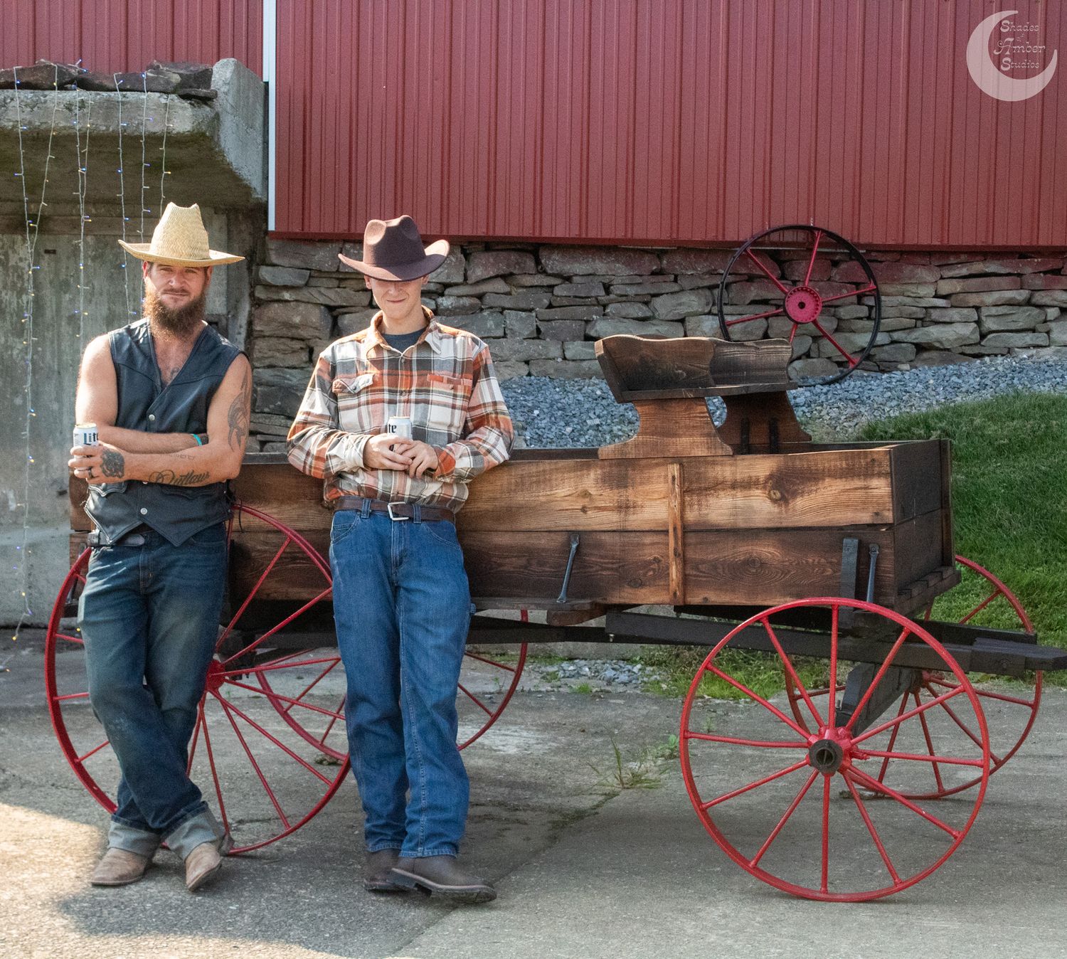 Two people in country attire leaning against antique wagon with red barn backdrop.