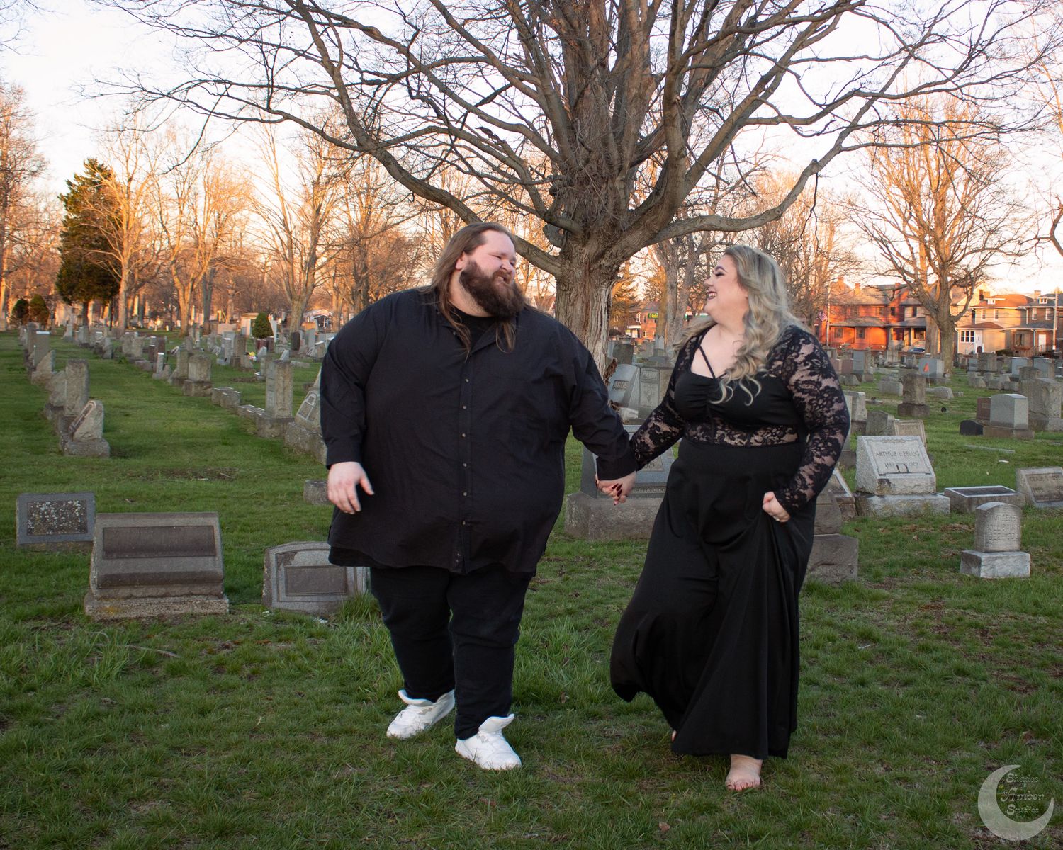 Couple in black attire holding hands in a cemetery at golden hour with gravestones and bare trees.