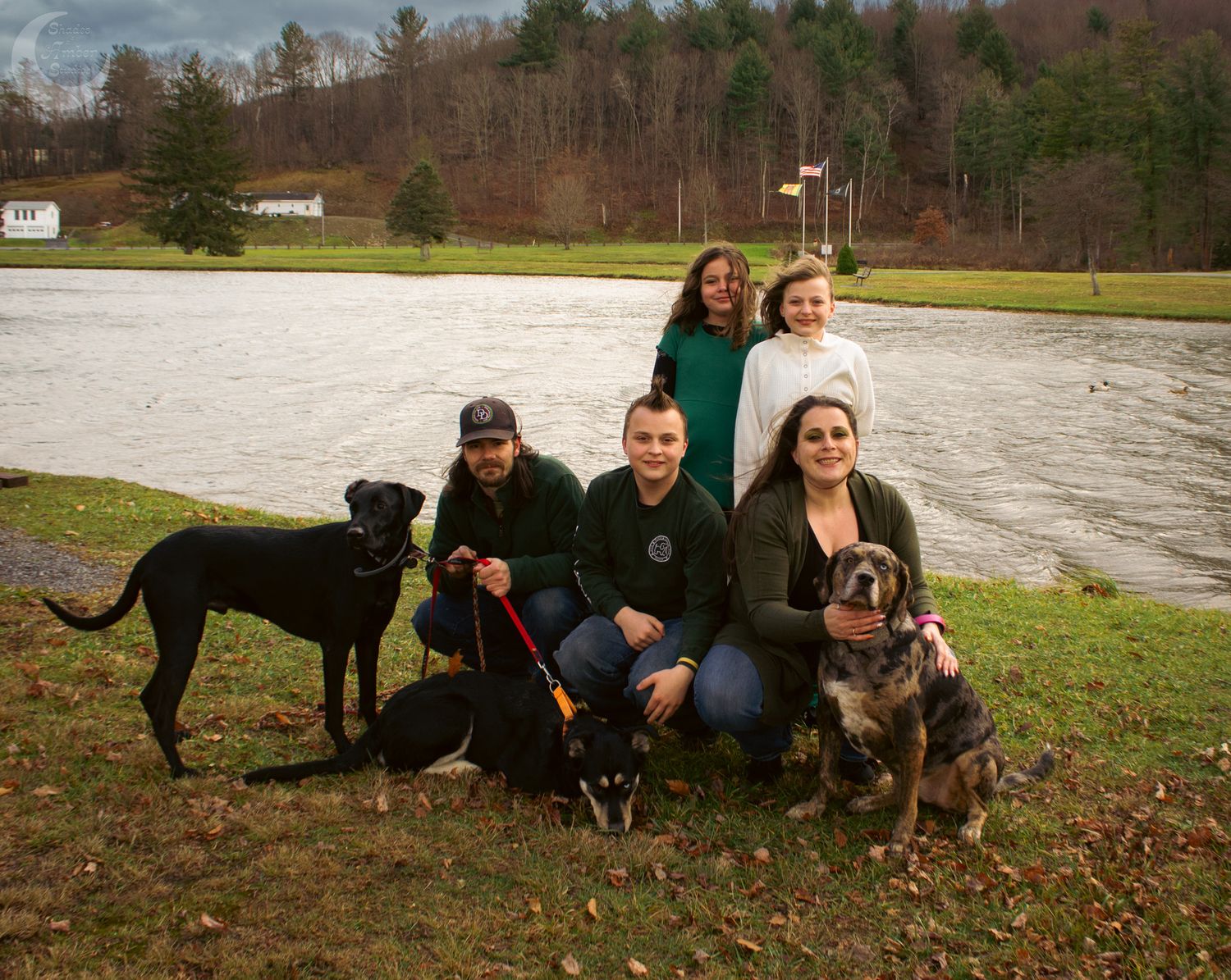 A group of people and their black dog pose together outdoors on a fall day in a grassy area.