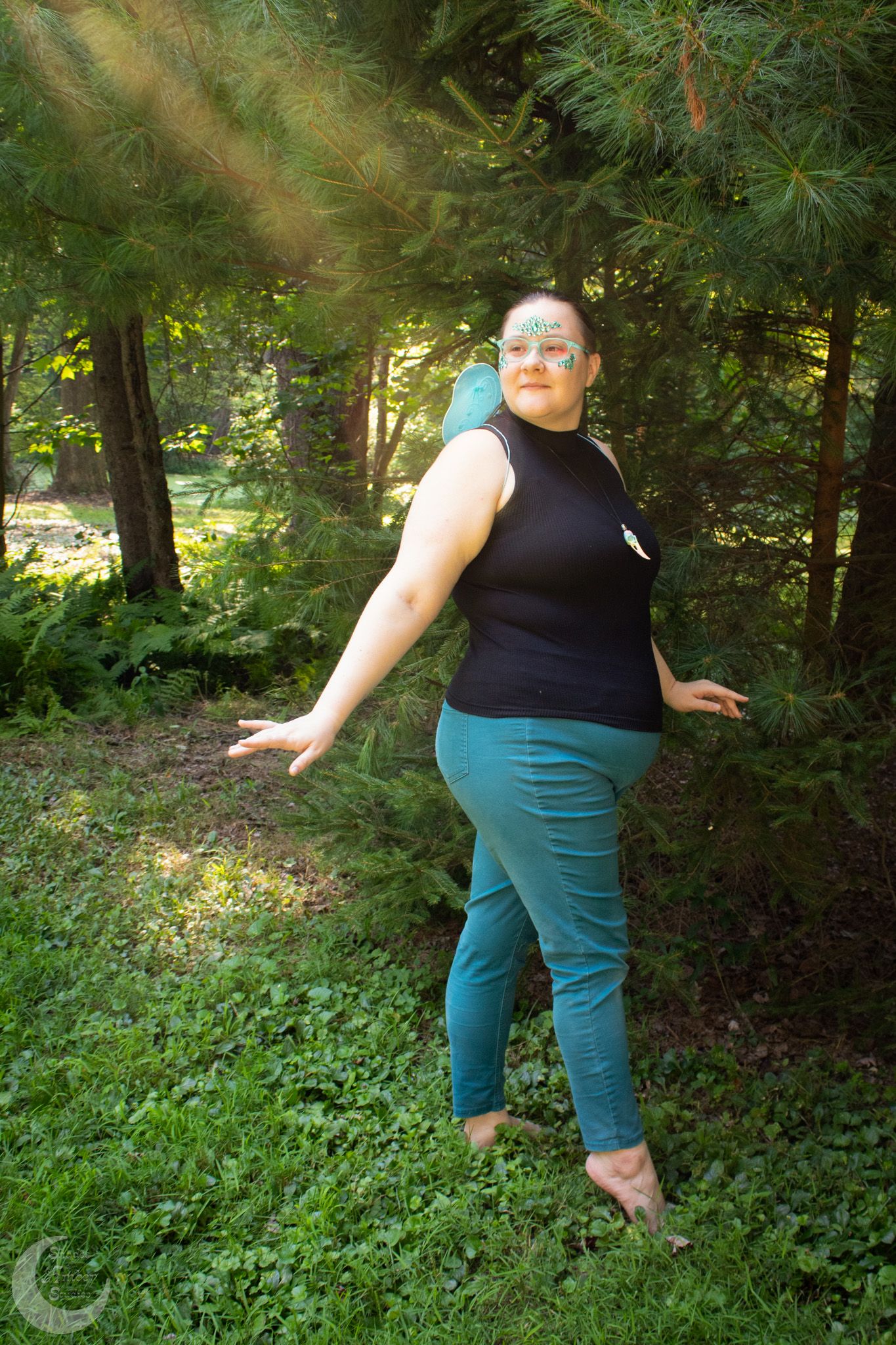 Figure in black tank top and teal capri pants dances playfully among pine trees in a sunlit forest setting.