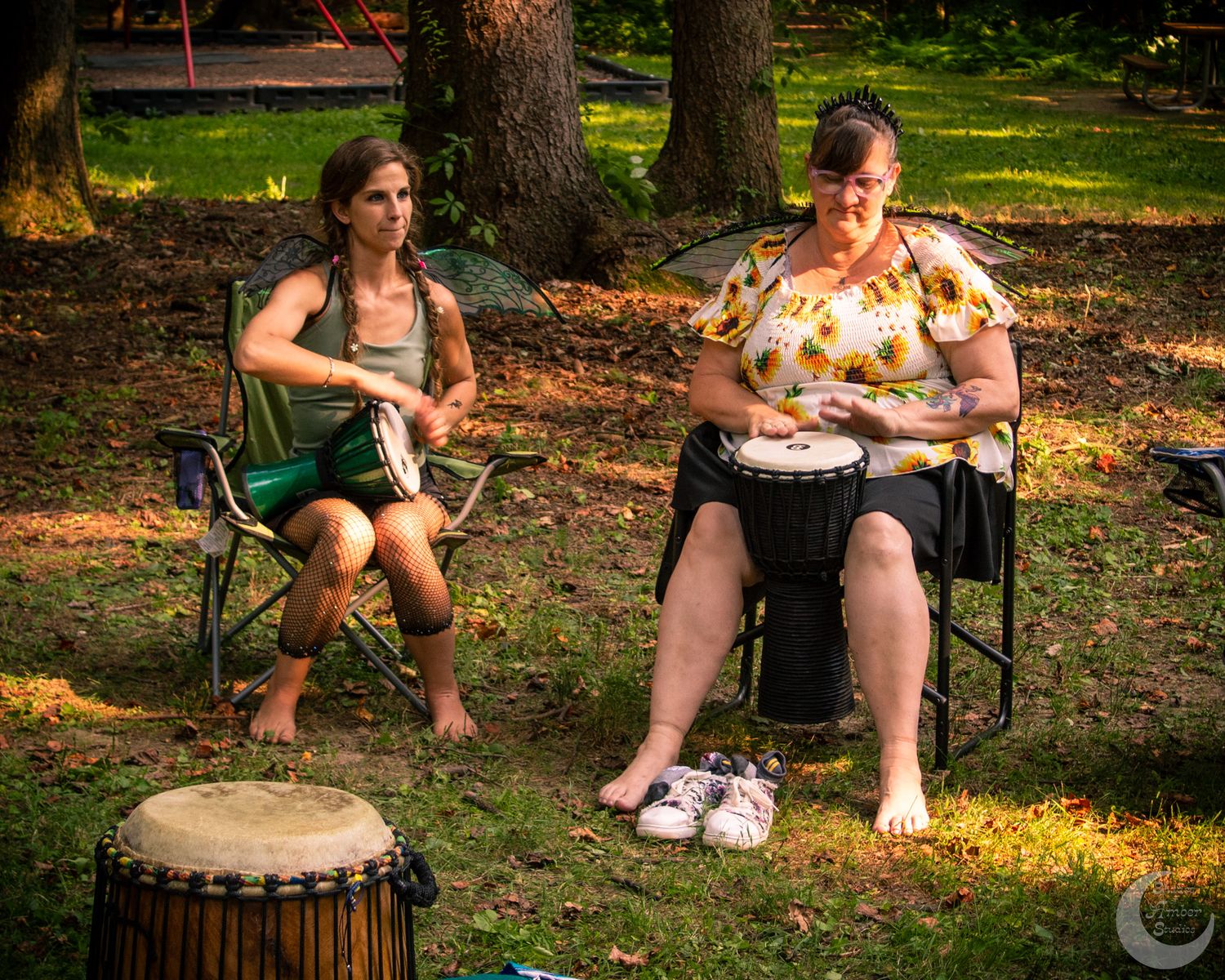 Two musicians play hand drums outdoors in a forest setting while sitting in camping chairs at dusk.