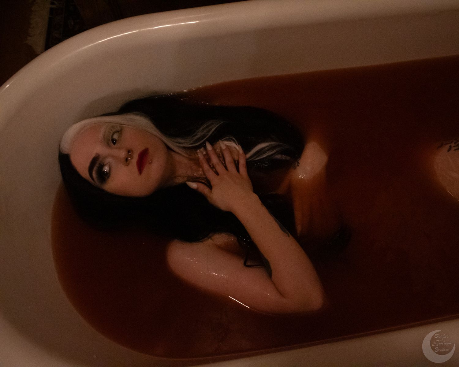 A woman with long dark hair reclines in a clawfoot bathtub filled with dark reddish-brown water.