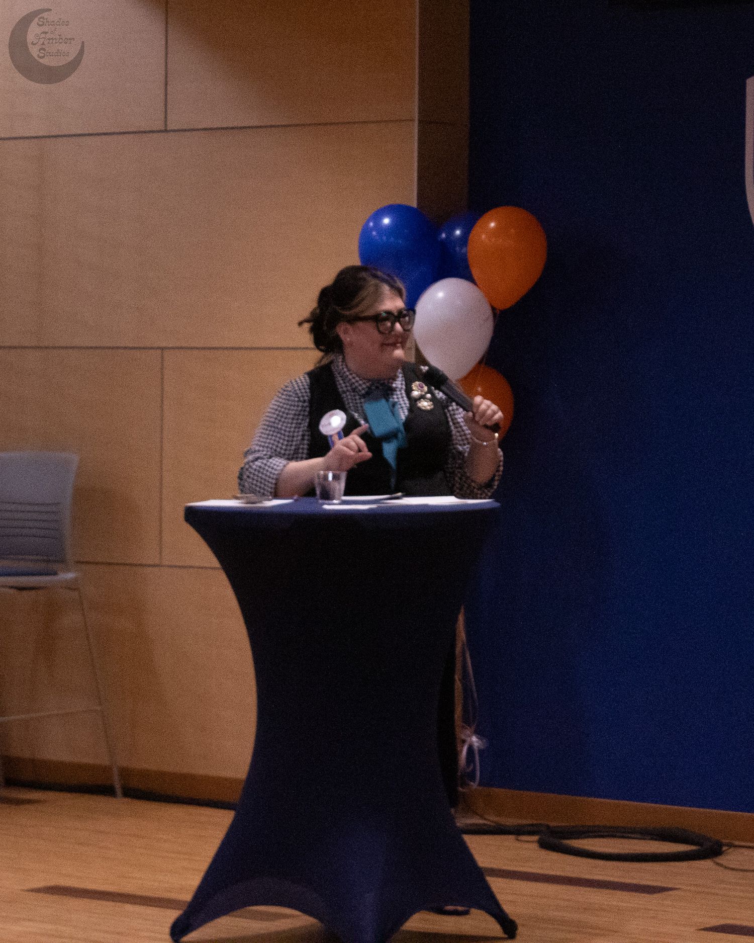 A speaker presents from behind a black podium stand with blue, orange and white balloons in the background.