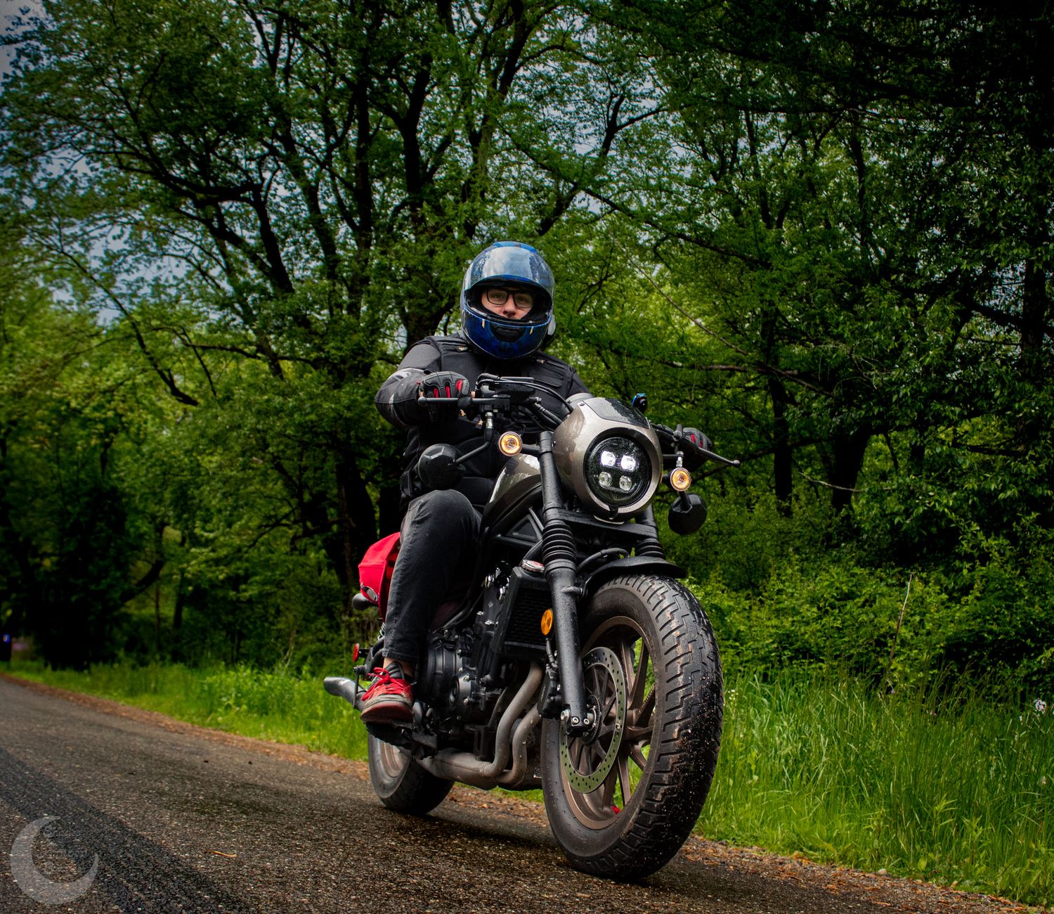 A motorcyclist rides a custom cafe racer style motorcycle on a tree-lined rural road on a sunny day.