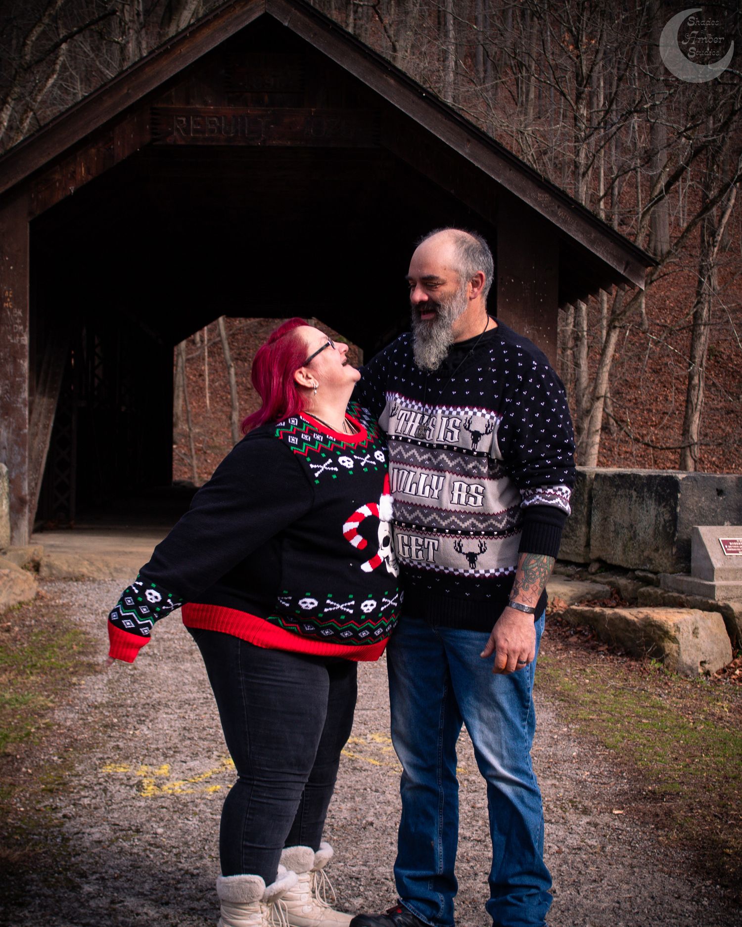 A couple wearing matching holiday sweaters share a tender moment under a rustic wooden shelter.