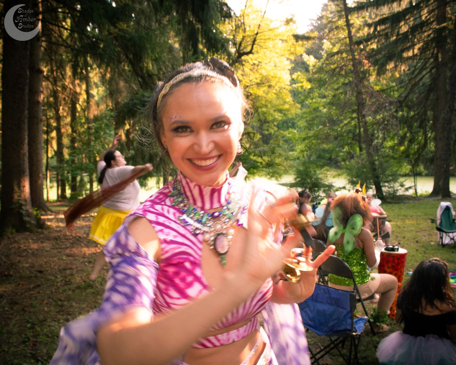 A person in a pink and white tie-dye shirt poses playfully in a park with lush greenery in the background.